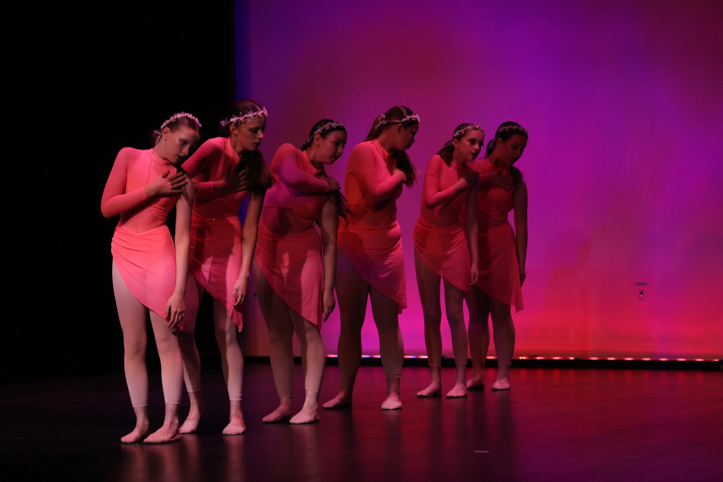 Six young ballet dancers in pink costumes standing in a line with their hands on their chests, bowing during a performance on stage with purple and pink lighting.