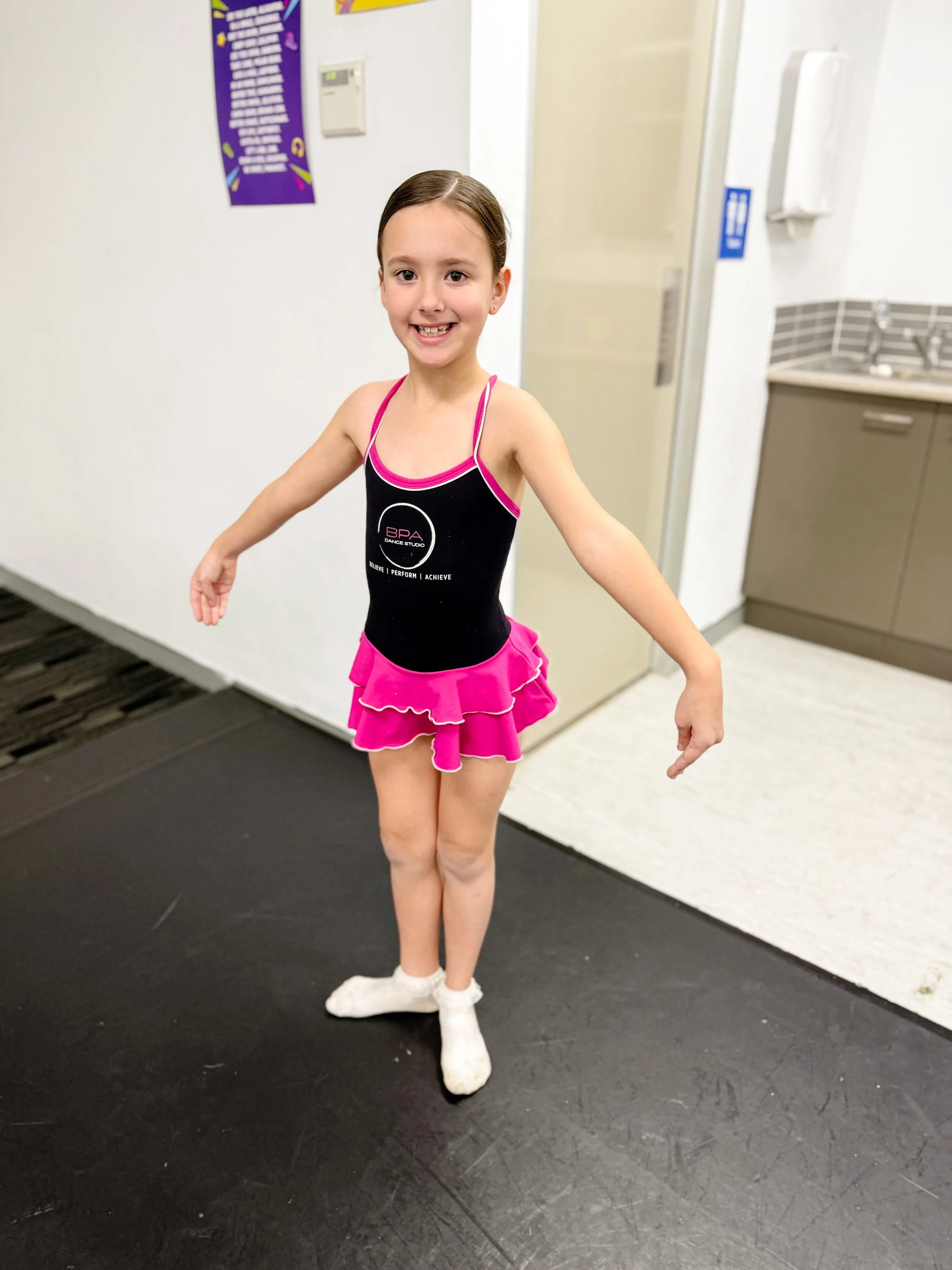 A young girl in a black and pink dance costume standing on a black floor, smiling with her arms out to her sides, in an indoor setting near a white wall, a door, and a kitchenette.
