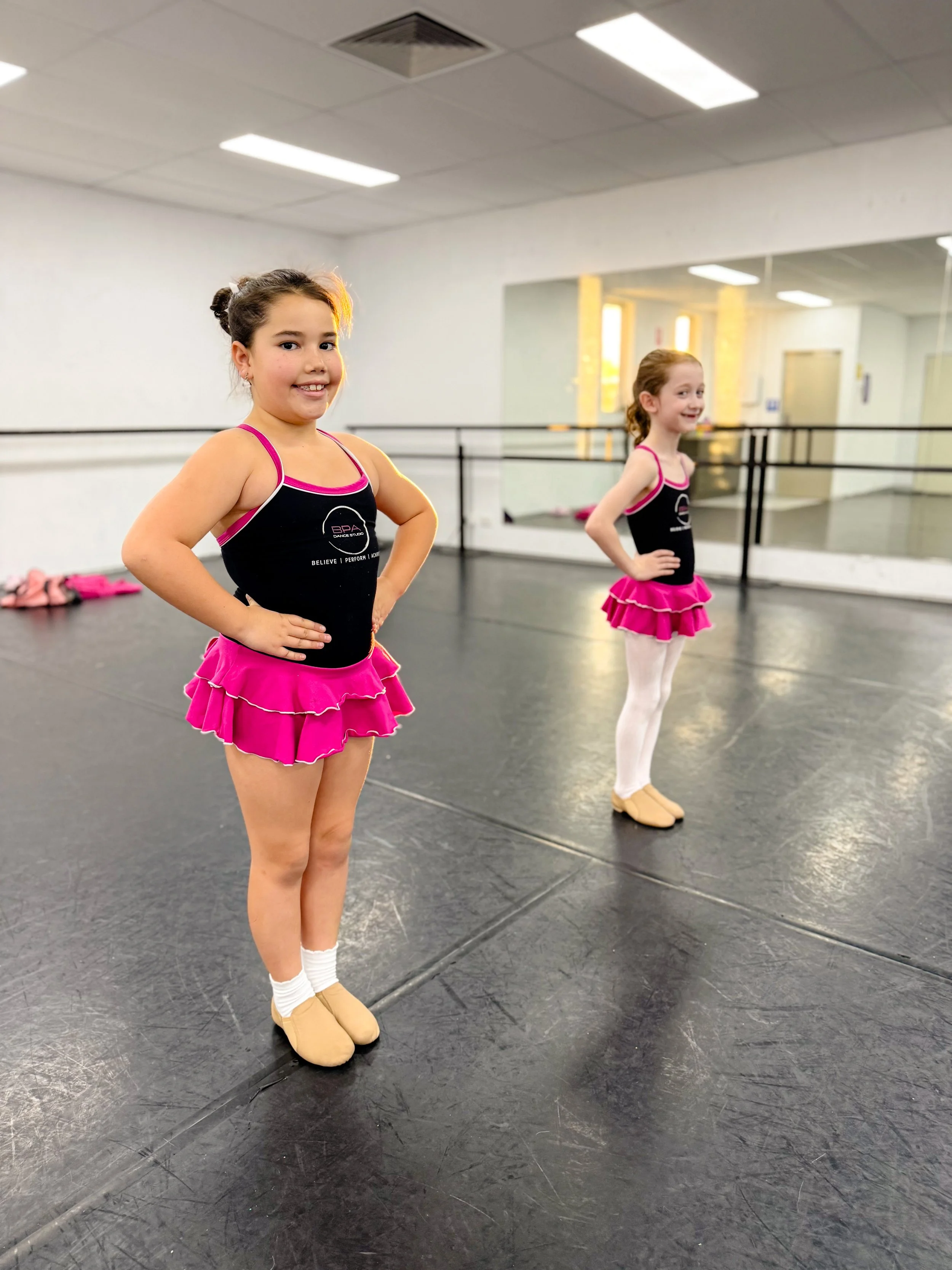 Two young girls in ballet costumes standing in front of a mirror in a dance studio, with hands on hips and smiling.