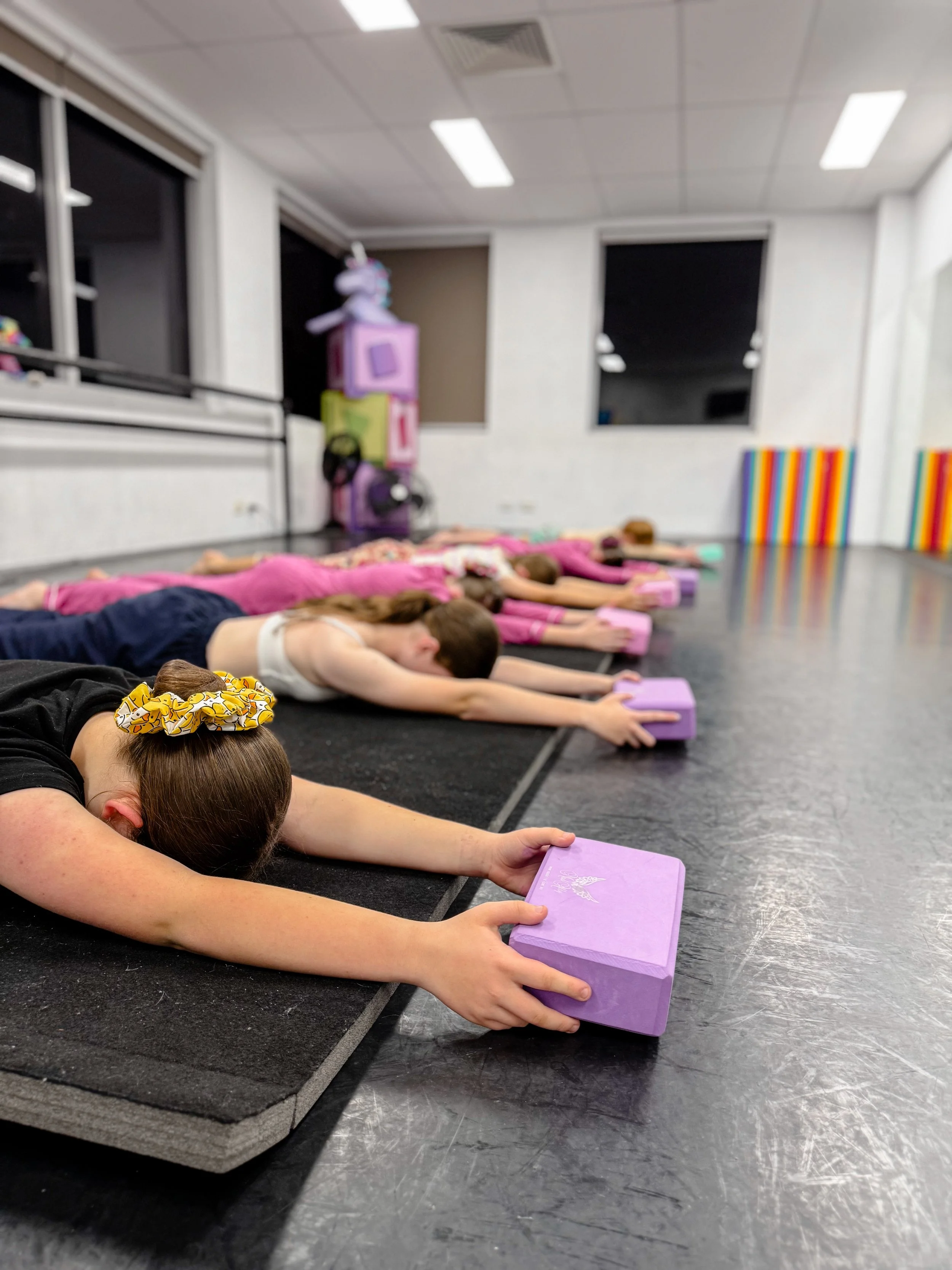 Young girls practicing yoga on the floor using purple blocks in a studio with black flooring, white walls, and large windows.