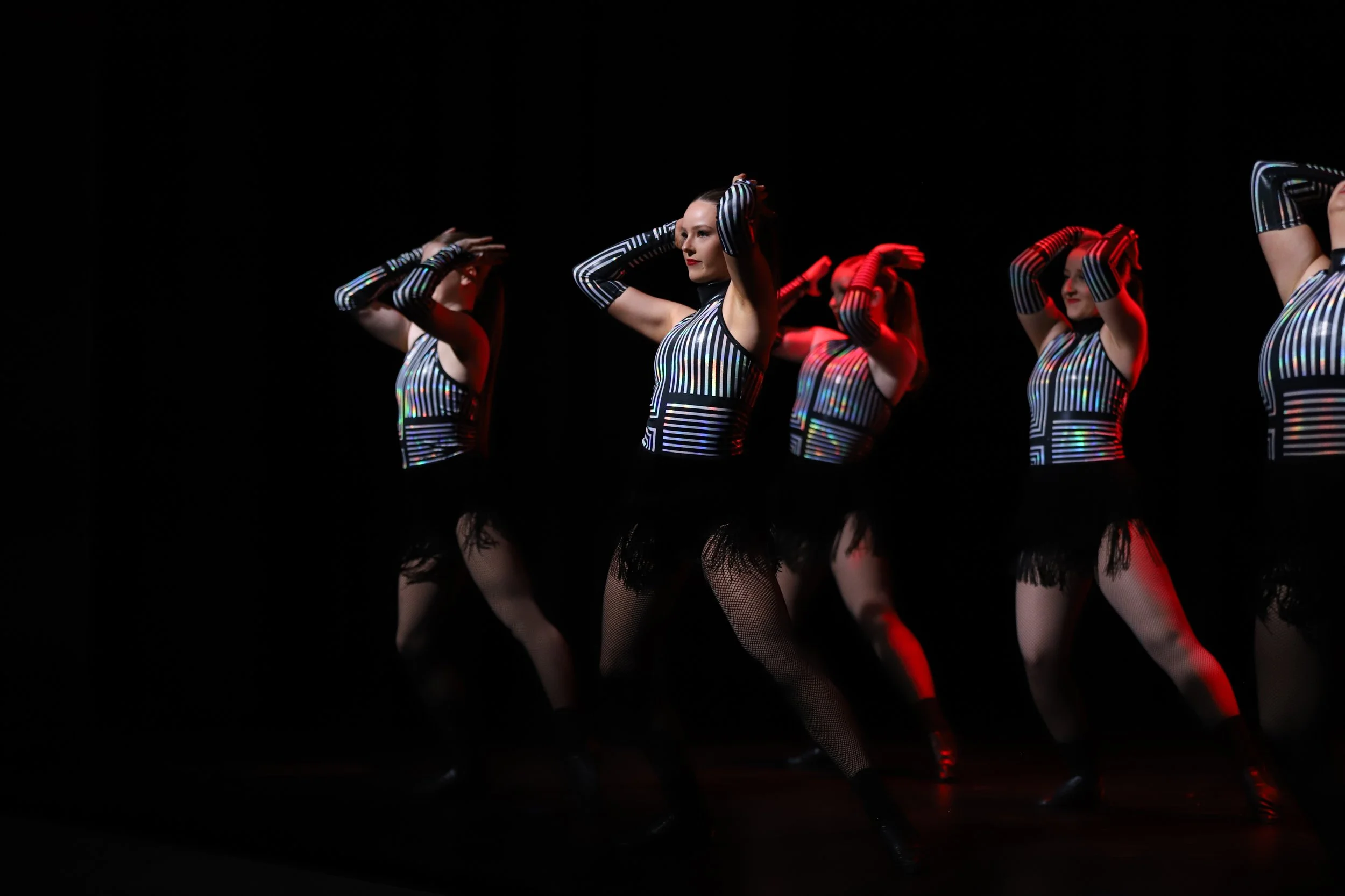 Group of women in matching futuristic striped costumes performing a dance on stage with dark background.