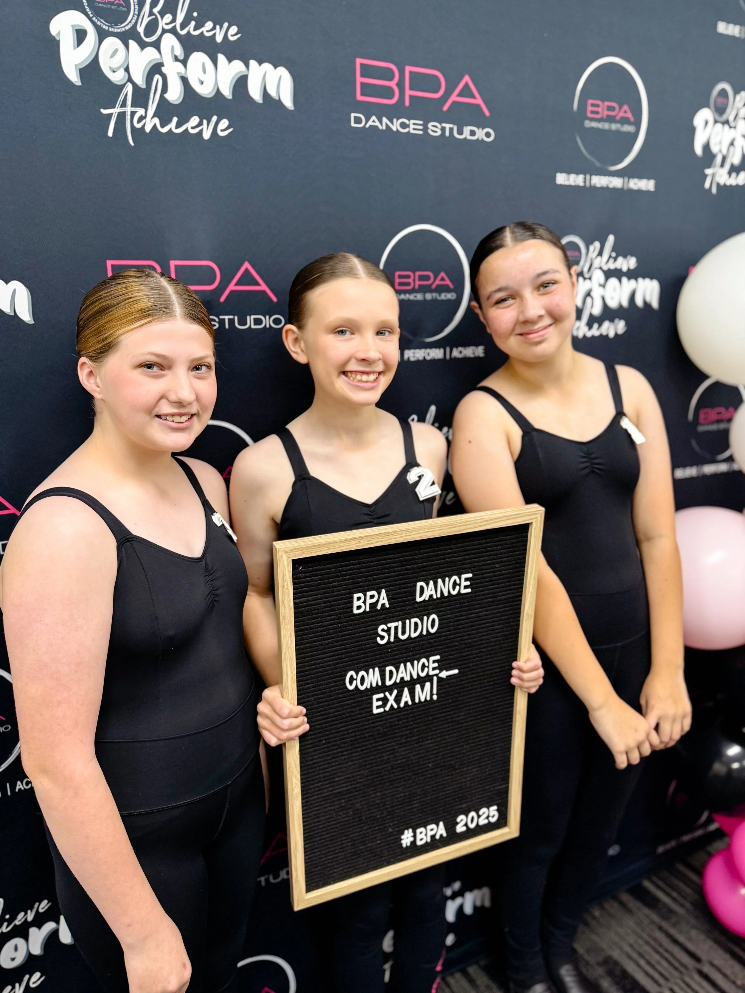 Three young girls dressed in black dance outfits standing in front of a backdrop with the BPA Dance Studio logo, holding a sign that reads 'BPA Dance Studio COM DANCE EXAM! #BPA 2025', smiling at the camera.