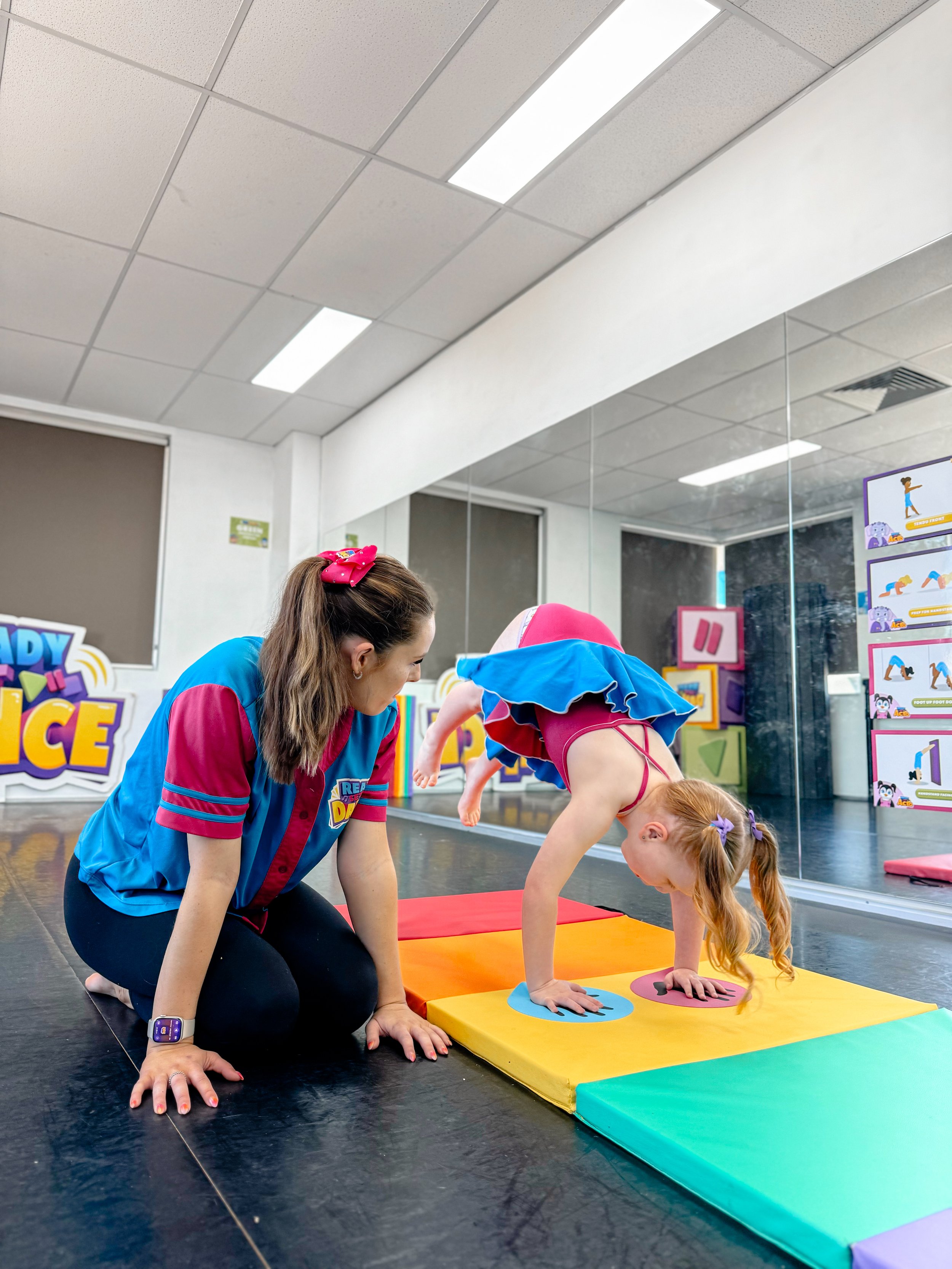 A young girl with red hair and ponytails doing a cartwheel on colorful gymnastics mats, assisted by an instructor in a blue and pink shirt in a dance or gymnastics studio with mirrors and educational posters on the wall.