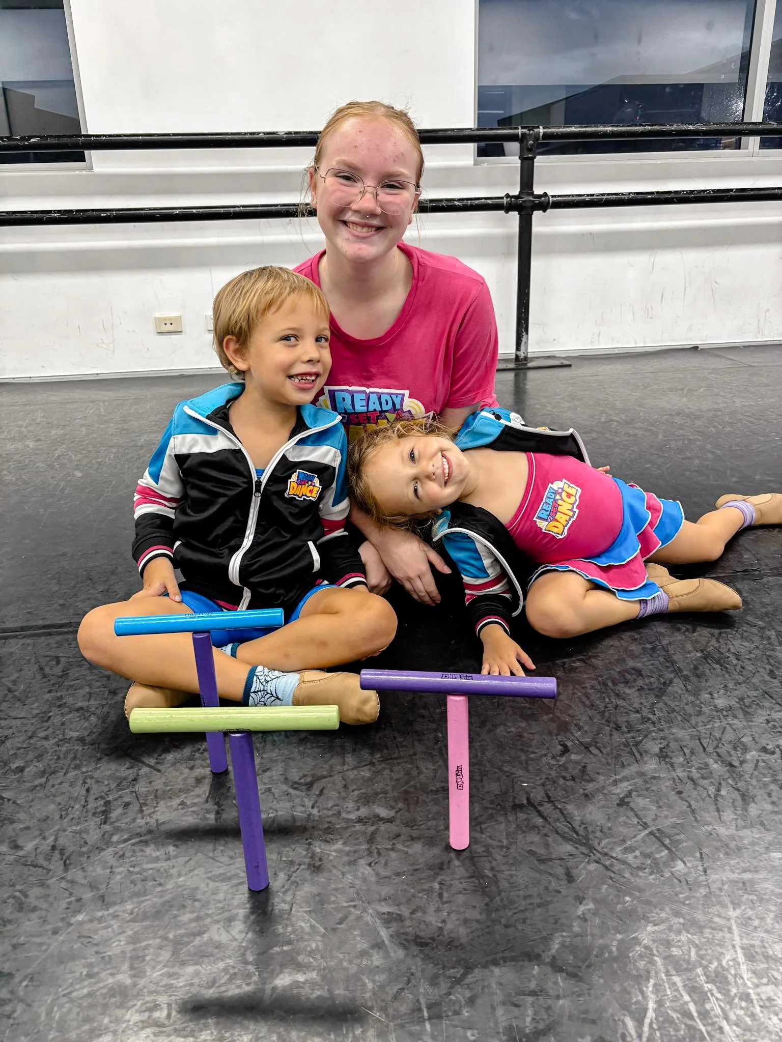 Three children in dance costumes and a young woman in a pink shirt sitting on the dance studio floor.