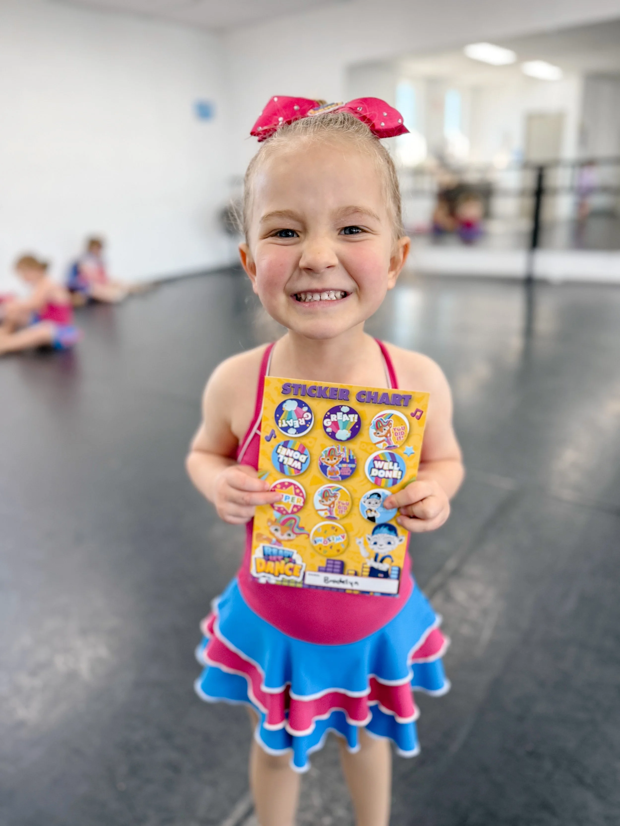 A young girl in a pink and blue dance costume smiling and holding a sticker chart at a dance studio.