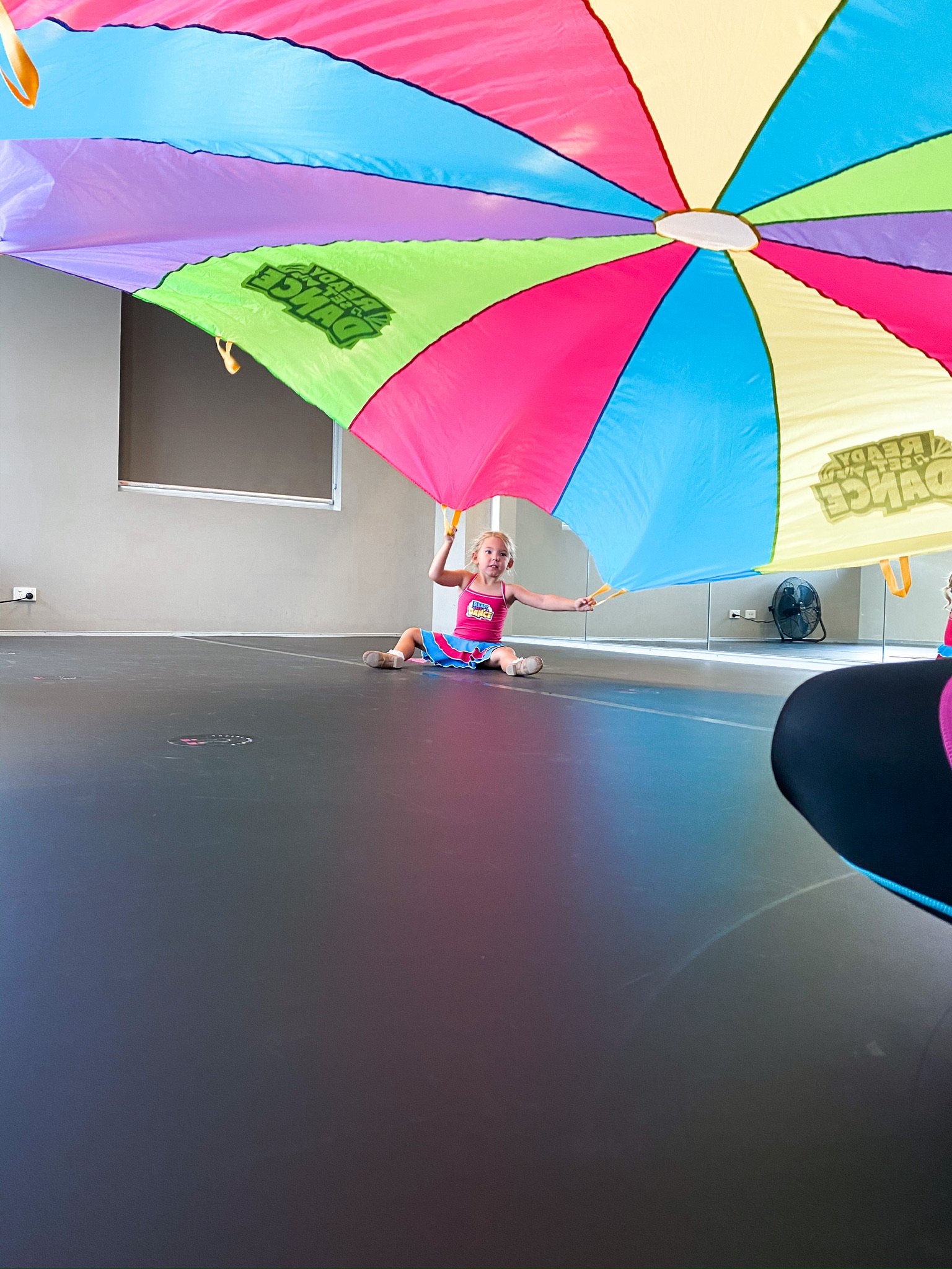 A young girl sitting on the floor indoors, holding a large colorful beach umbrella.