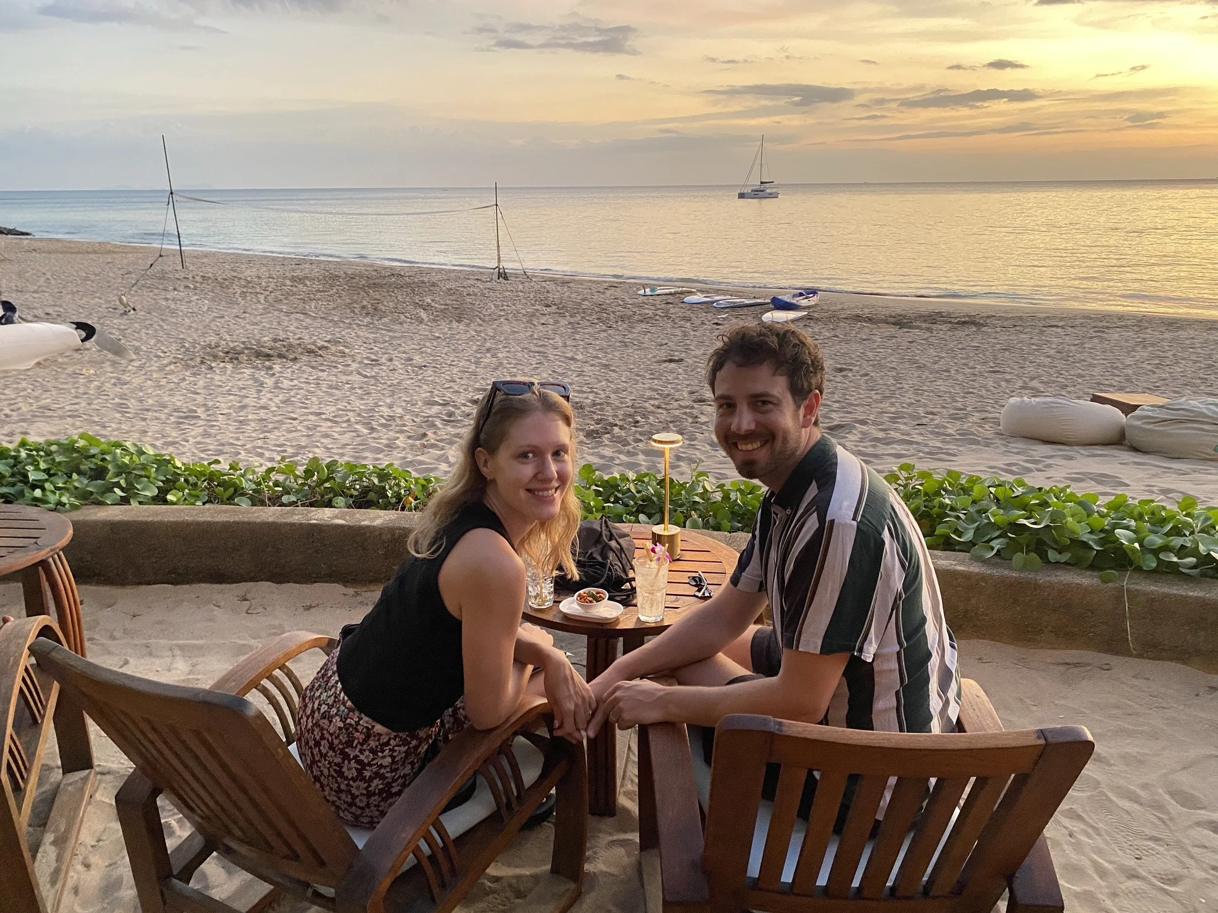 A smiling couple sitting at a table on a beach during sunset, with boats and the ocean in the background.