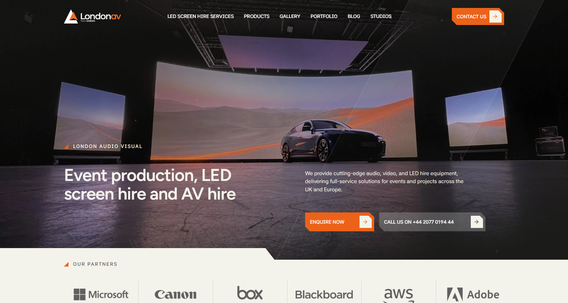 Website homepage for London AV, an event production company. The page features a large LED screen display showing a landscape scene with mountains and a sunset, and a black car parked in front. Navigation menu at the top includes options like LED Screen Hire Services, Products, Gallery, Portfolio, Blog, Studios. Contact button at the top right and an enquiry button at the bottom left are visible. Logos of partners such as Microsoft, Canon, Box, Blackboard, AWS, and Adobe are shown at the bottom.