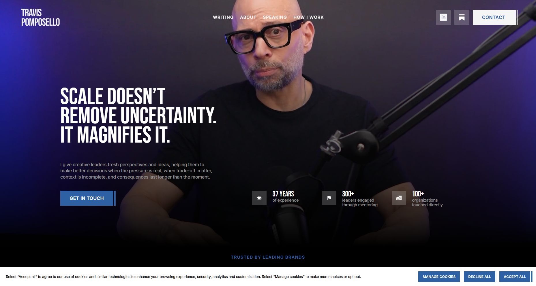 A man wearing glasses with a beard, sitting in front of a microphone, on a dark purple background with blue lighting, on a website homepage for Travis Pomposello, a creative professional.
