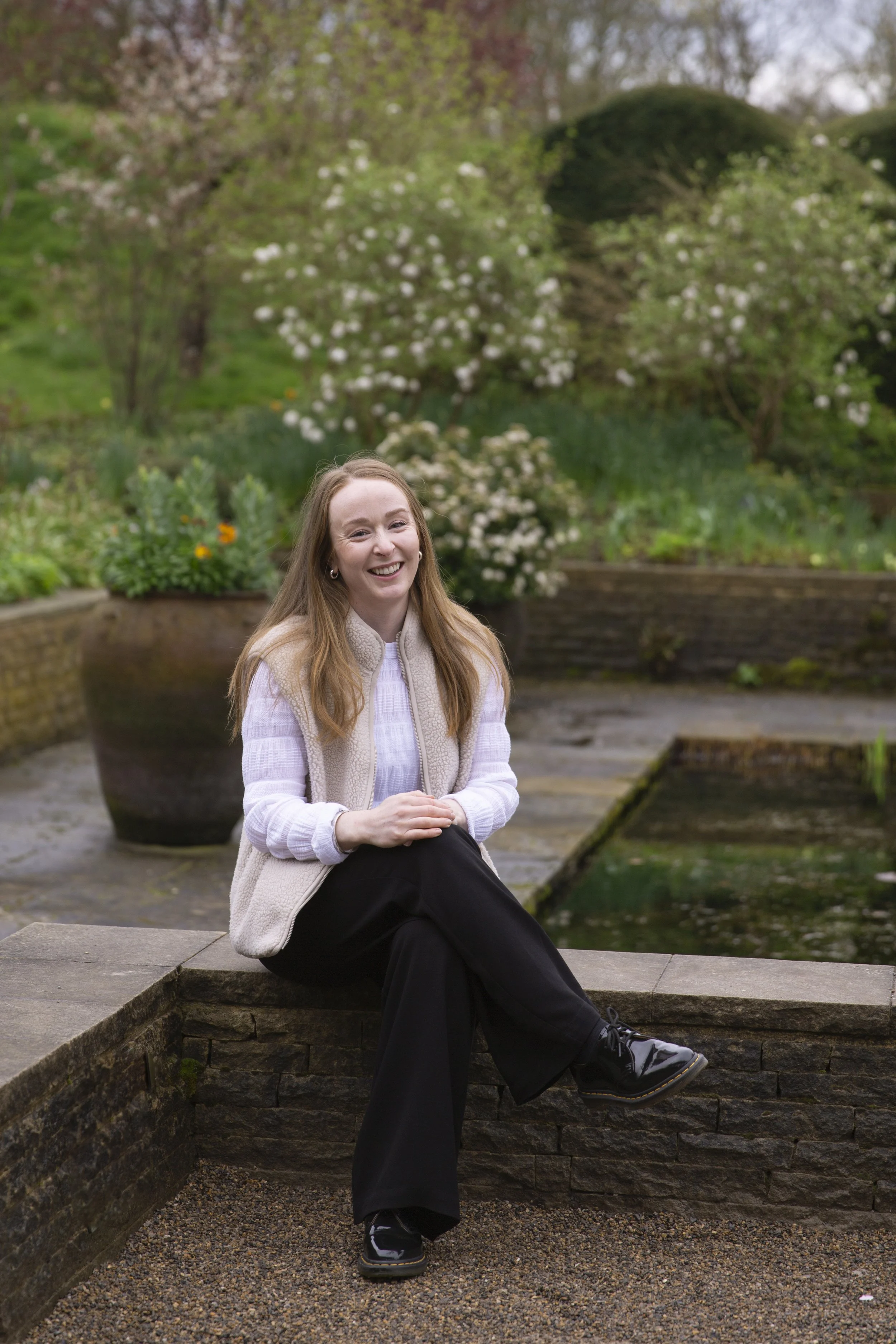 A woman sitting on a low stone wall by a garden pond, smiling and looking at the camera, with flowering trees and lush greenery in the background.