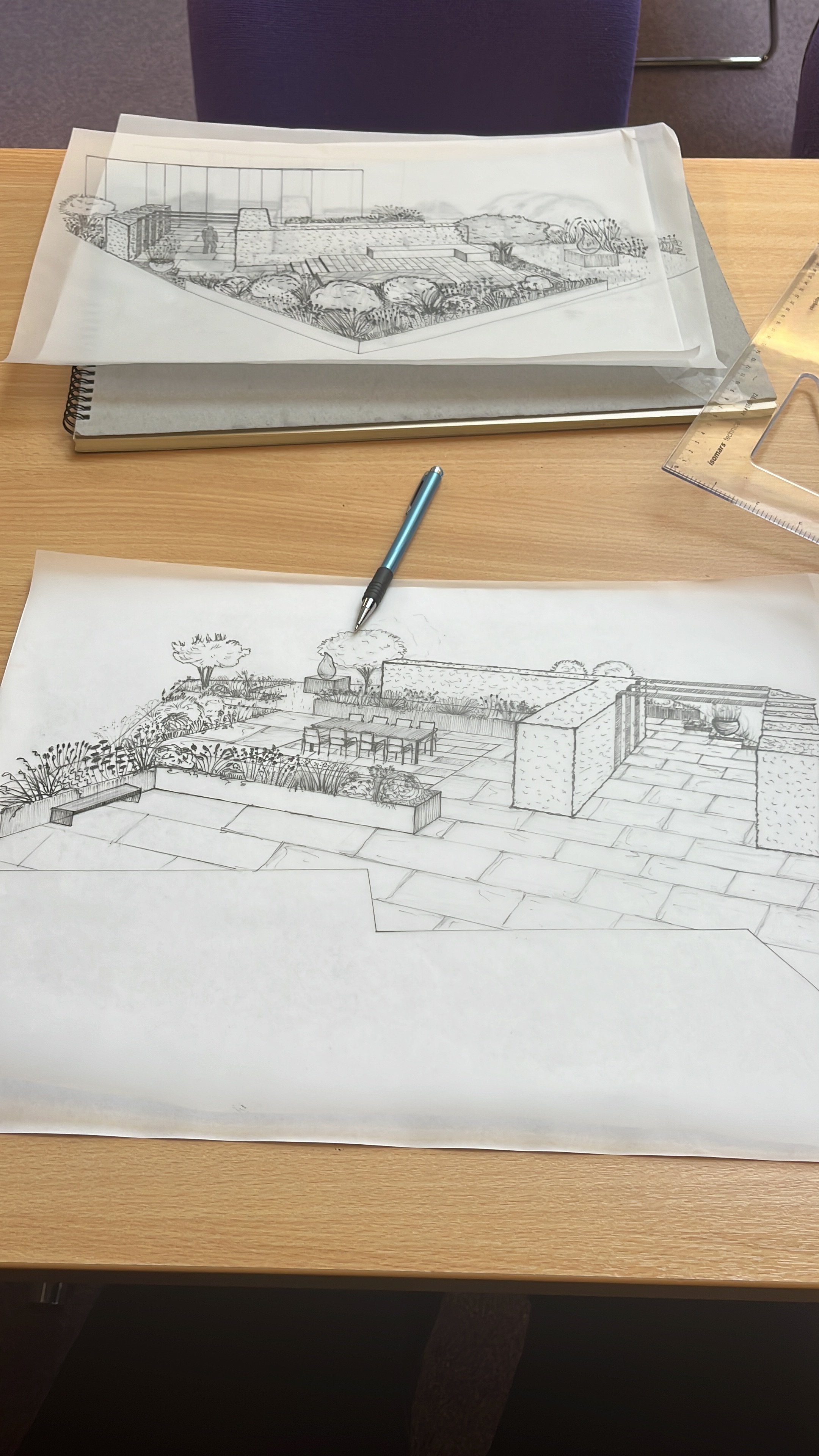 A detailed pencil sketch of a landscaped outdoor area featuring a patio, garden beds with various plants, and a bench, with a ruler and pen nearby.