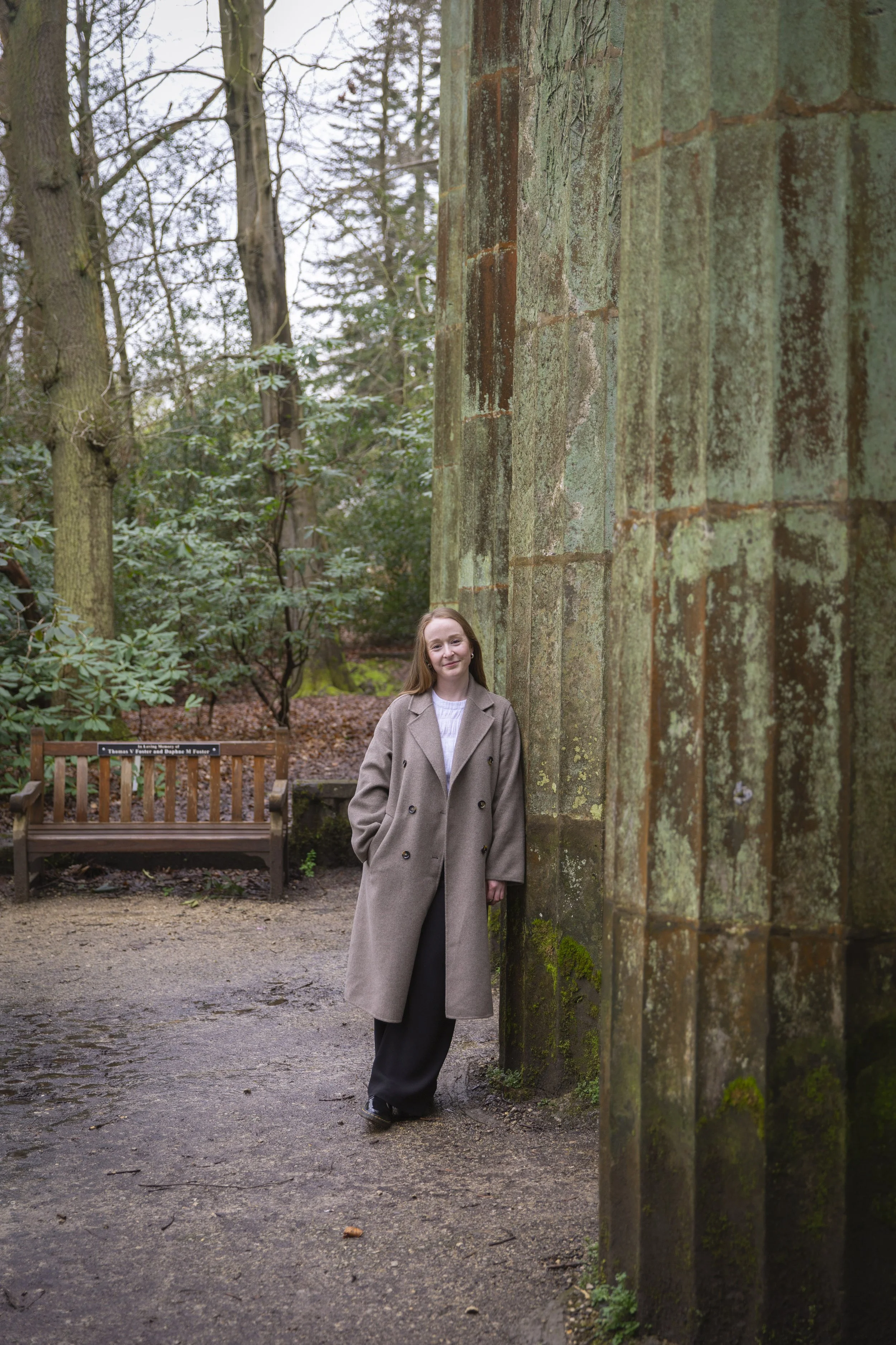A young woman with long hair standing outdoors in a wooded area, leaning against a moss-covered stone pillar, dressed in a beige coat and black pants, with a wooden bench and trees in the background.