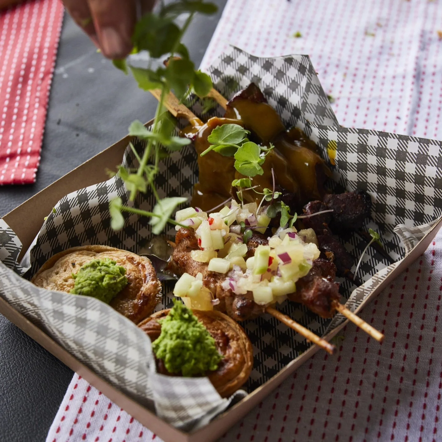A takeout box with grilled meat skewers topped with chopped onions, two potato salads with green guacamole, and microgreens, on a red and white checkered tablecloth.