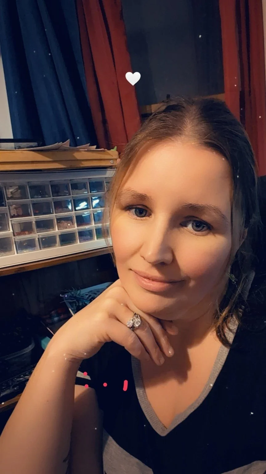 Close-up of a woman with brown hair and blue eyes, smiling, resting her chin on her hand with a large ring, in a room with dark blue and red curtains and a storage box in the background.