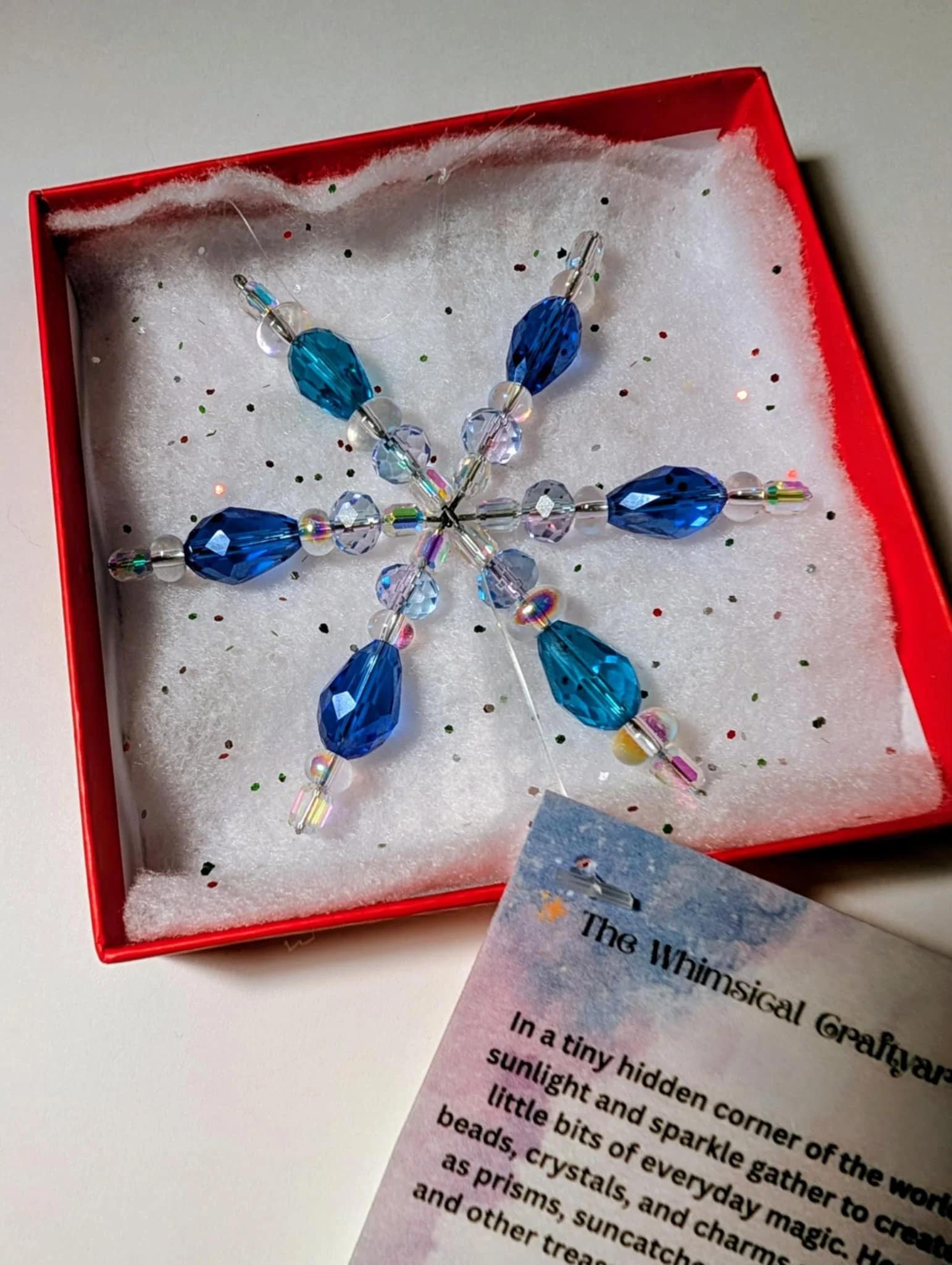 A snowflake-shaped decoration made of blue and clear beads in a red box, with a colorful info card partially visible.