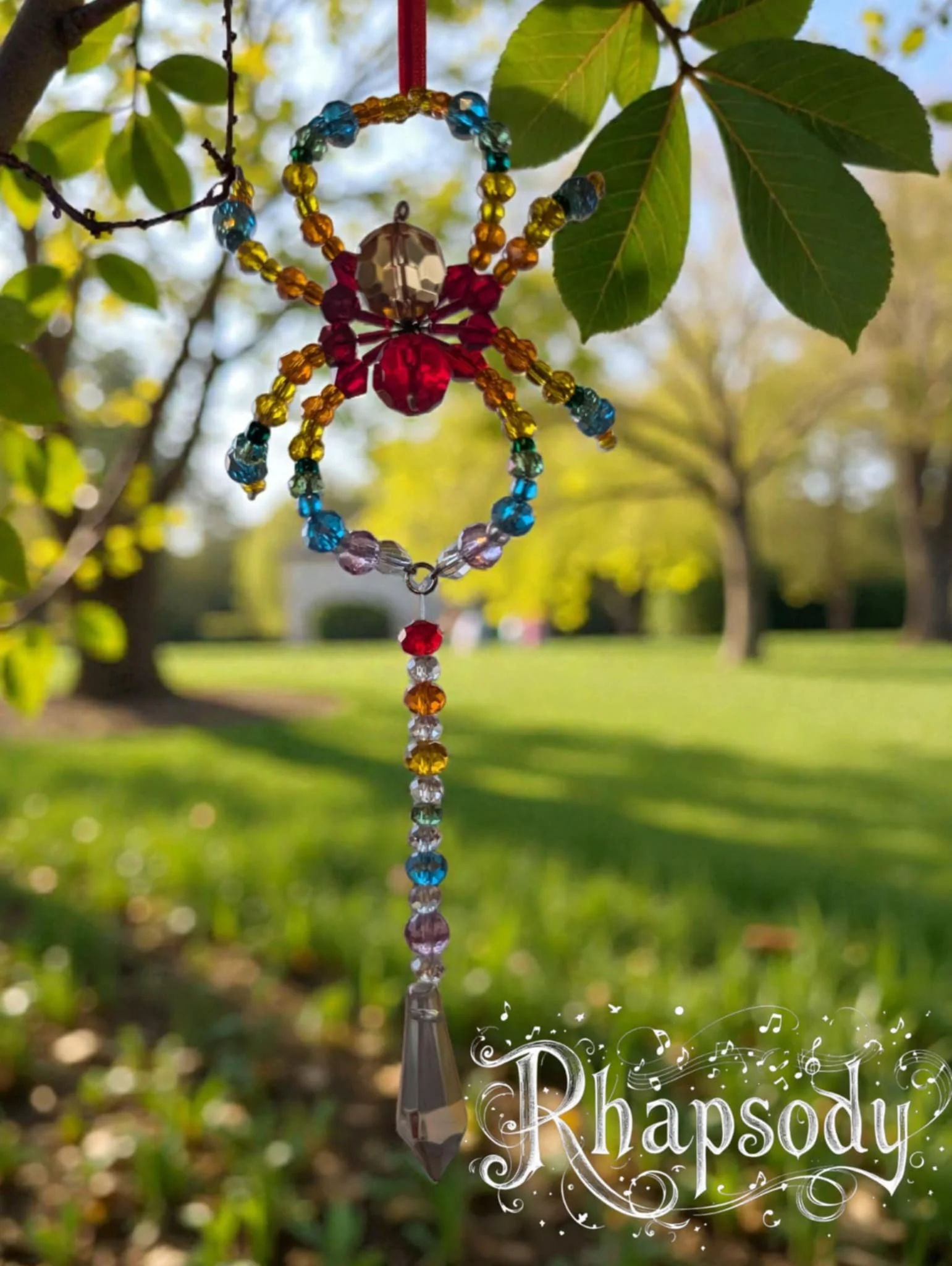 Colorful beaded butterfly wind chime hanging from a tree branch in a sunny park with green leaves and grass.