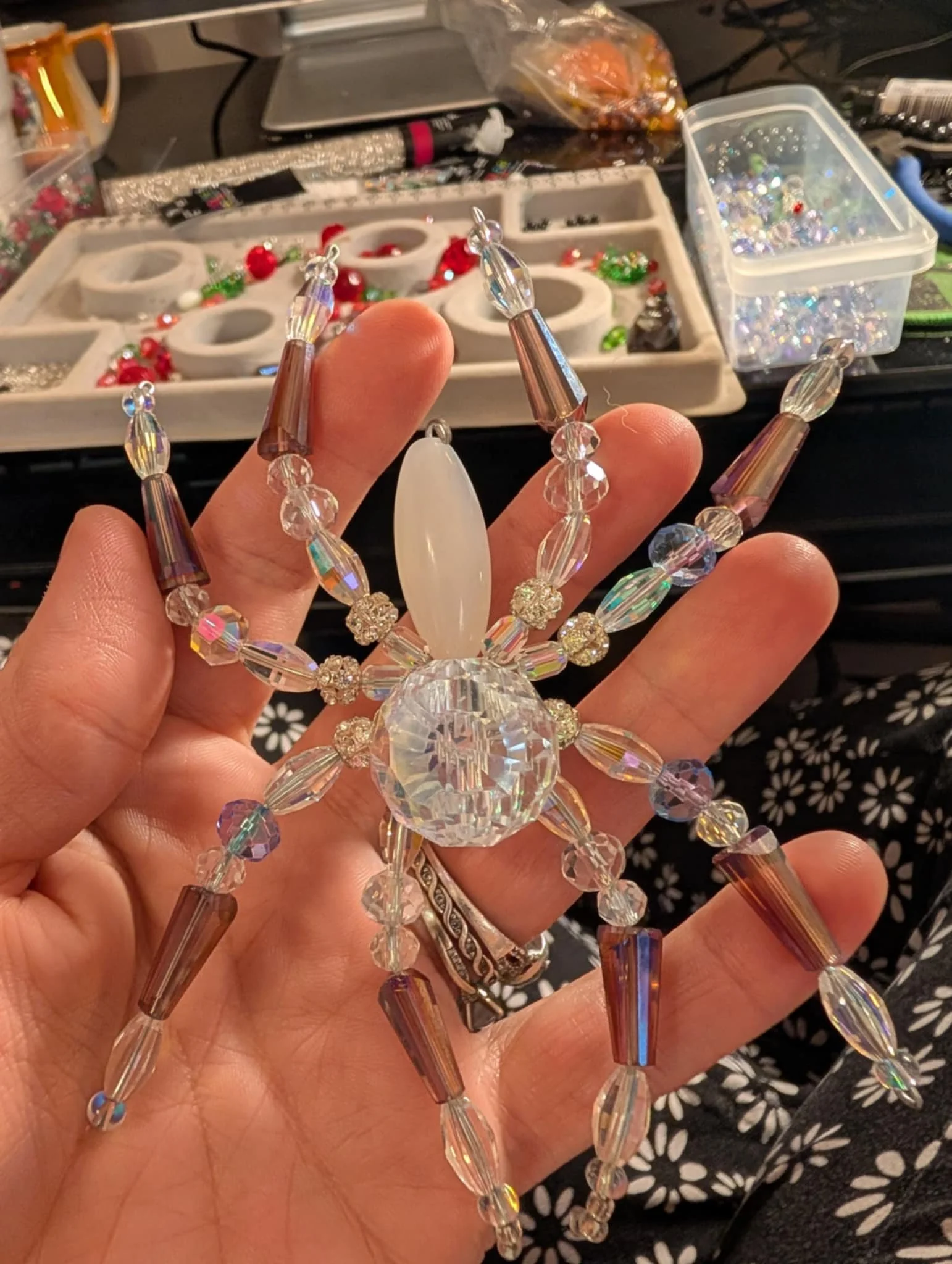 A sparkling, decorative crab-shaped ornament made of clear and iridescent beads, held in a person's hand with a jewelry-making workspace in the background.