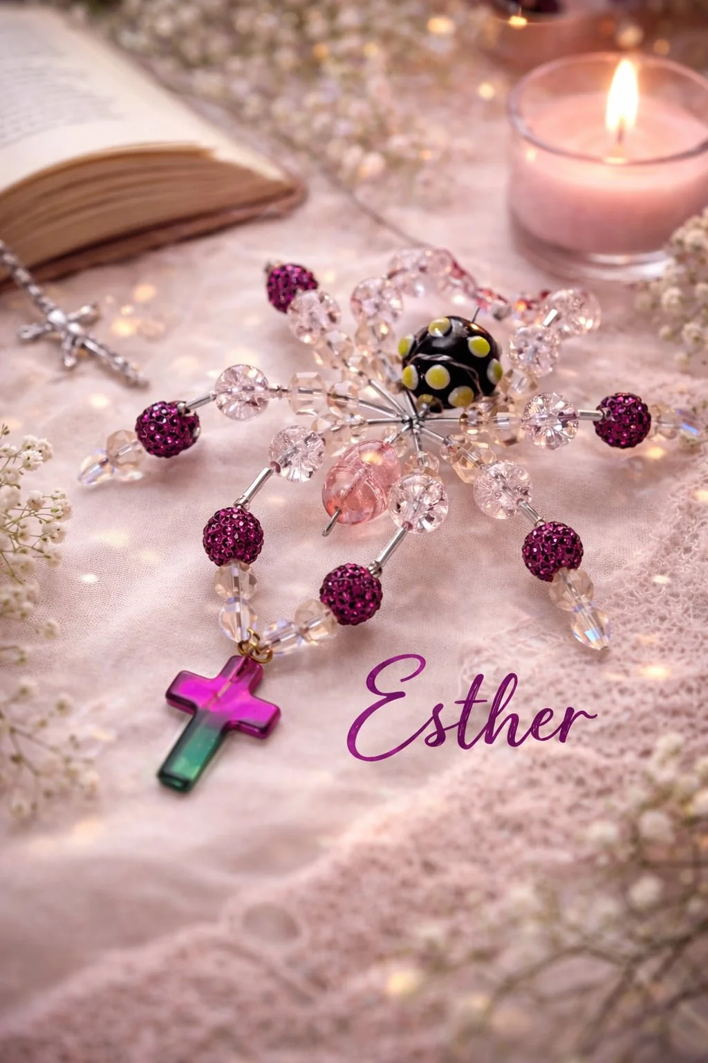 A rosary with pink, clear, and black beads, a pink and green cross, and a black bead with yellow dots, placed on a pink lace fabric with a lit candle, flowers, a cross necklace, an open book, and the name 'Esther' written in purple.