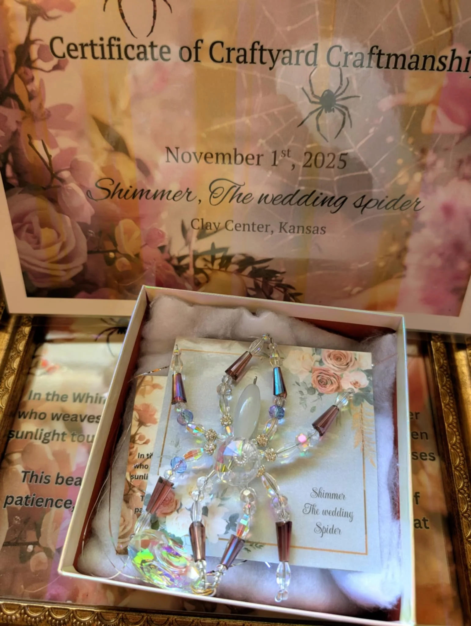 A decorative necklace with beads and crystals in a gift box, with a certificate of craftmanship in the background. The certificate indicates an event on November 1, 2025, at the Clay Center, Kansas, themed 'Shimmer, The Wedding Spider.'