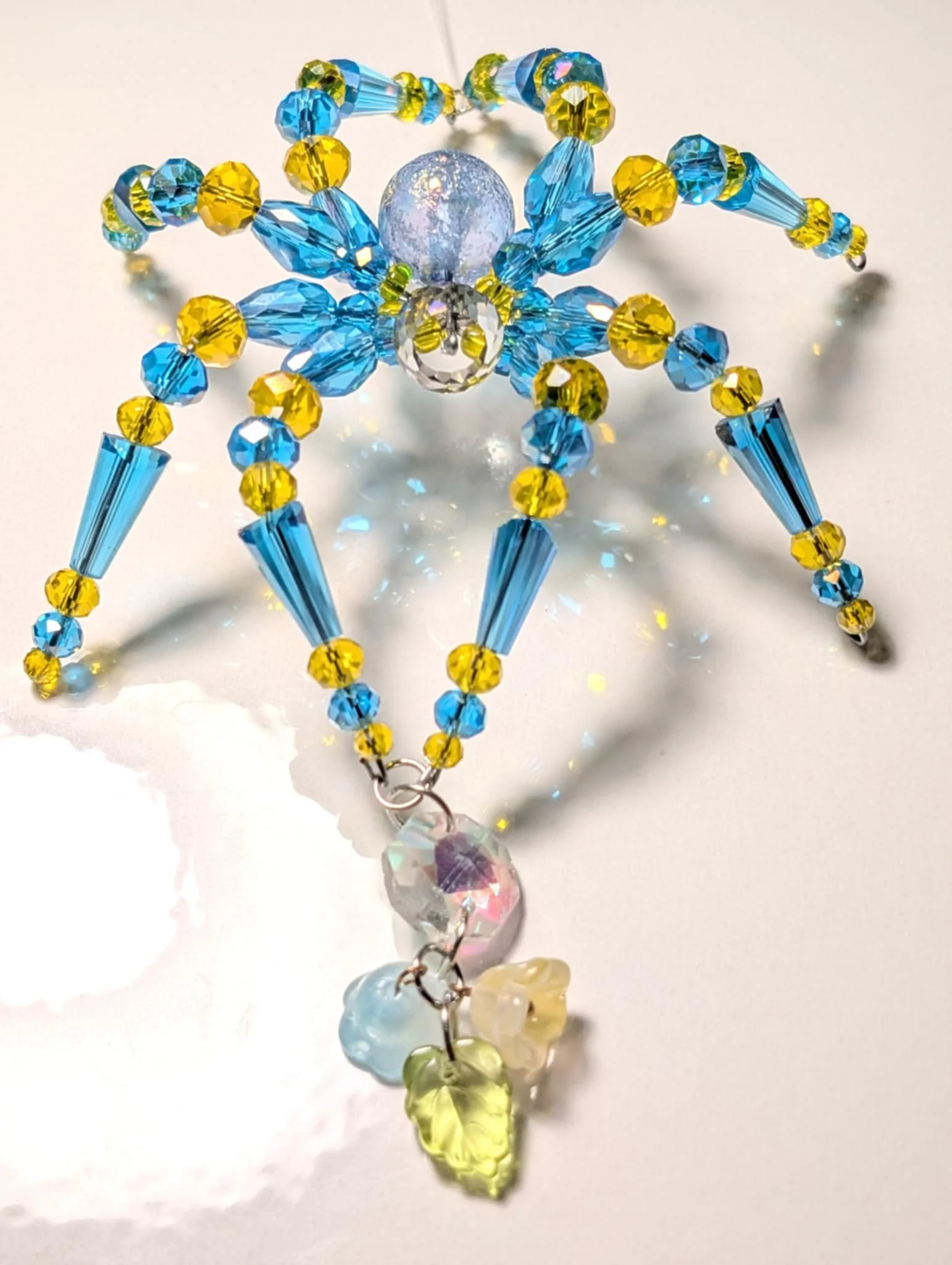 Colorful beaded angel ornament with blue, yellow, and clear beads, featuring a silver filigree bell and heart-shaped charms hanging from the bottom.