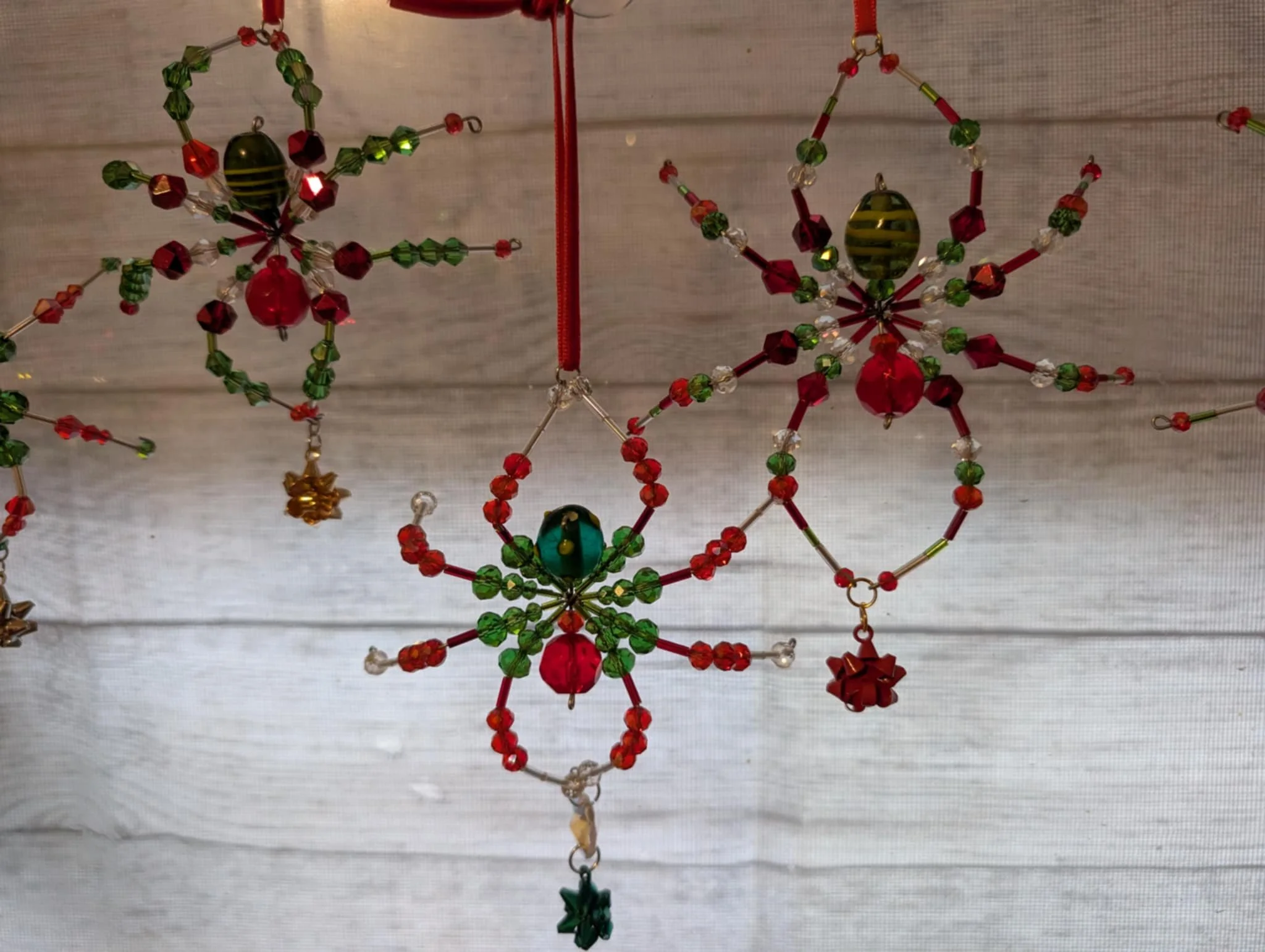 Holiday ornaments made of beads and wire in the shape of snowflakes with Christmas colors of red, green, and gold, hanging against a white wooden background.