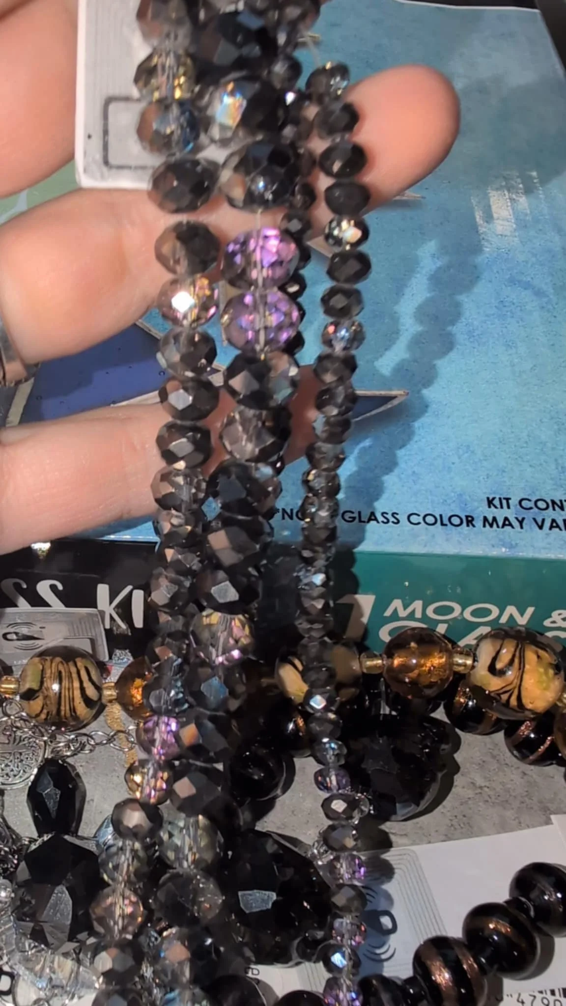 Close-up of a hand holding several strands of black and purple beads, possibly jewelry or decorative accessories, with a blurred background of jewelry and craft supplies.