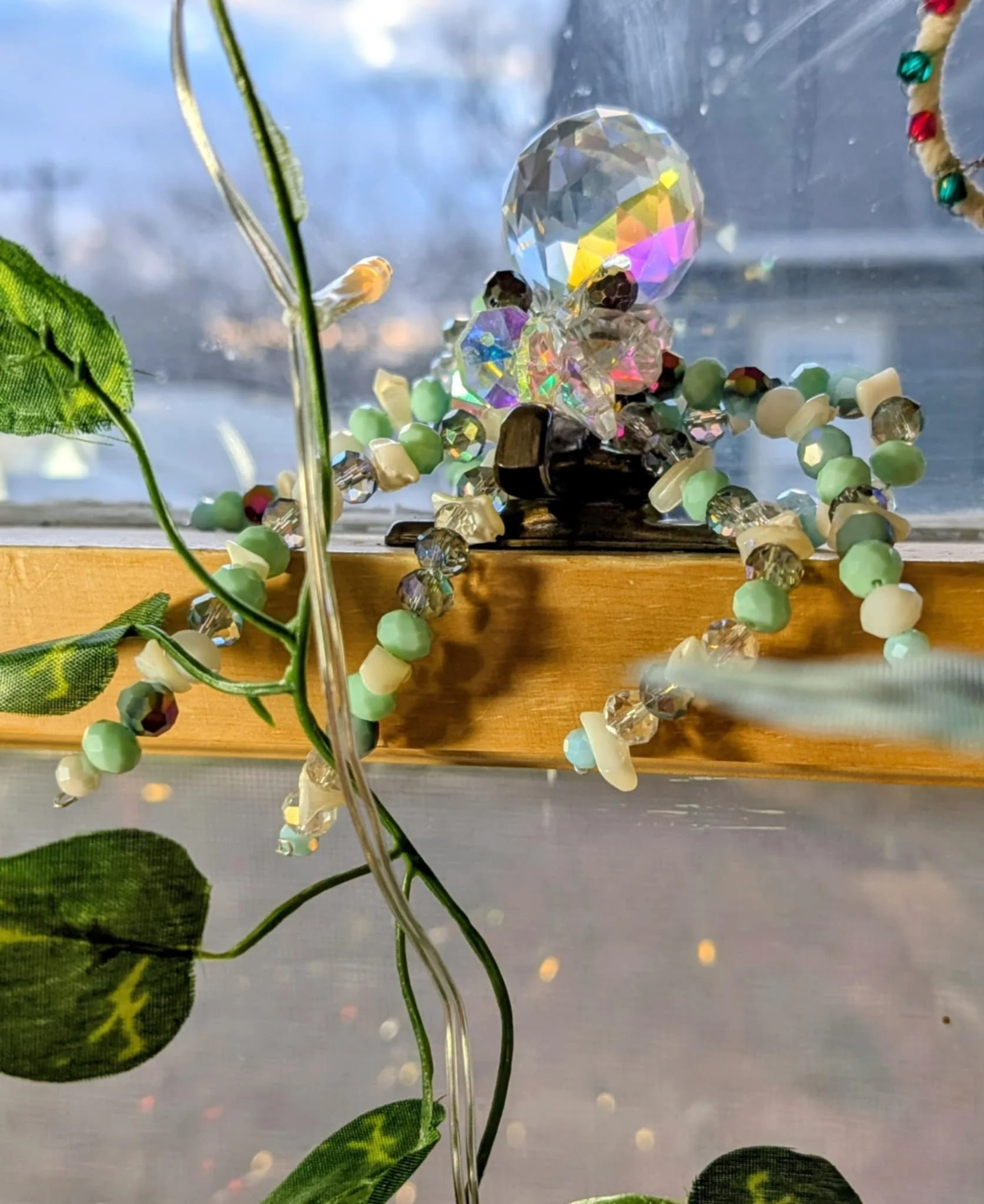 Decorative angel figurine made of colorful beads and crystals, positioned on a wooden sill with a window and blue sky in the background. Green leaves and a string of fairy lights are nearby.