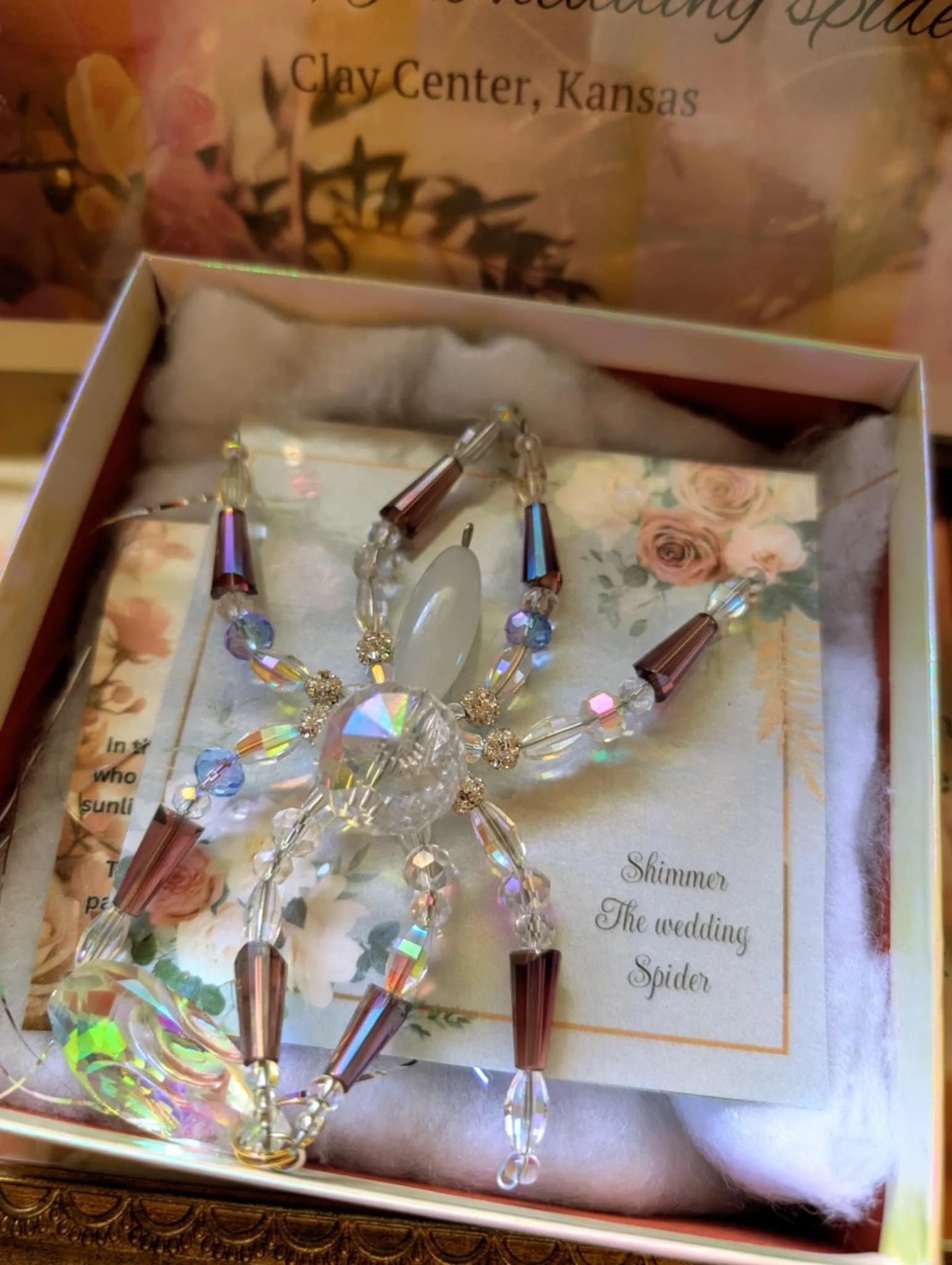 A sparkling, clear crystal wedding spider ornament with iridescent beads and metallic accents, resting on a floral decorated card inside a box.