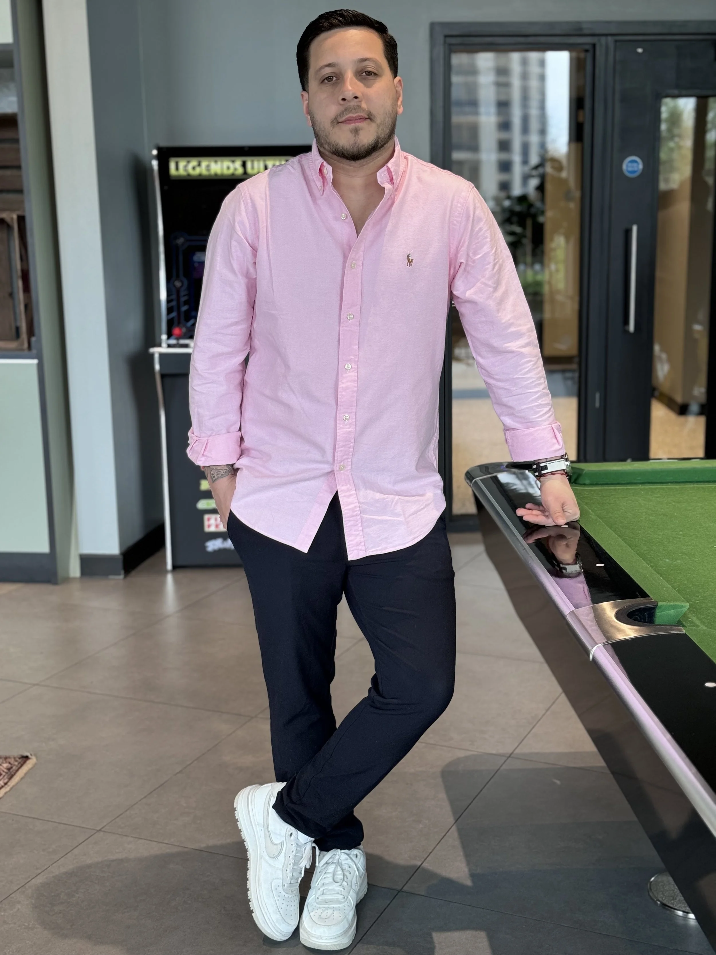 A man wearing a pink button-up shirt, dark pants, and white sneakers standing next to a pool table with his hand on the edge, in an indoor setting with glass doors and a pinball machine in the background.