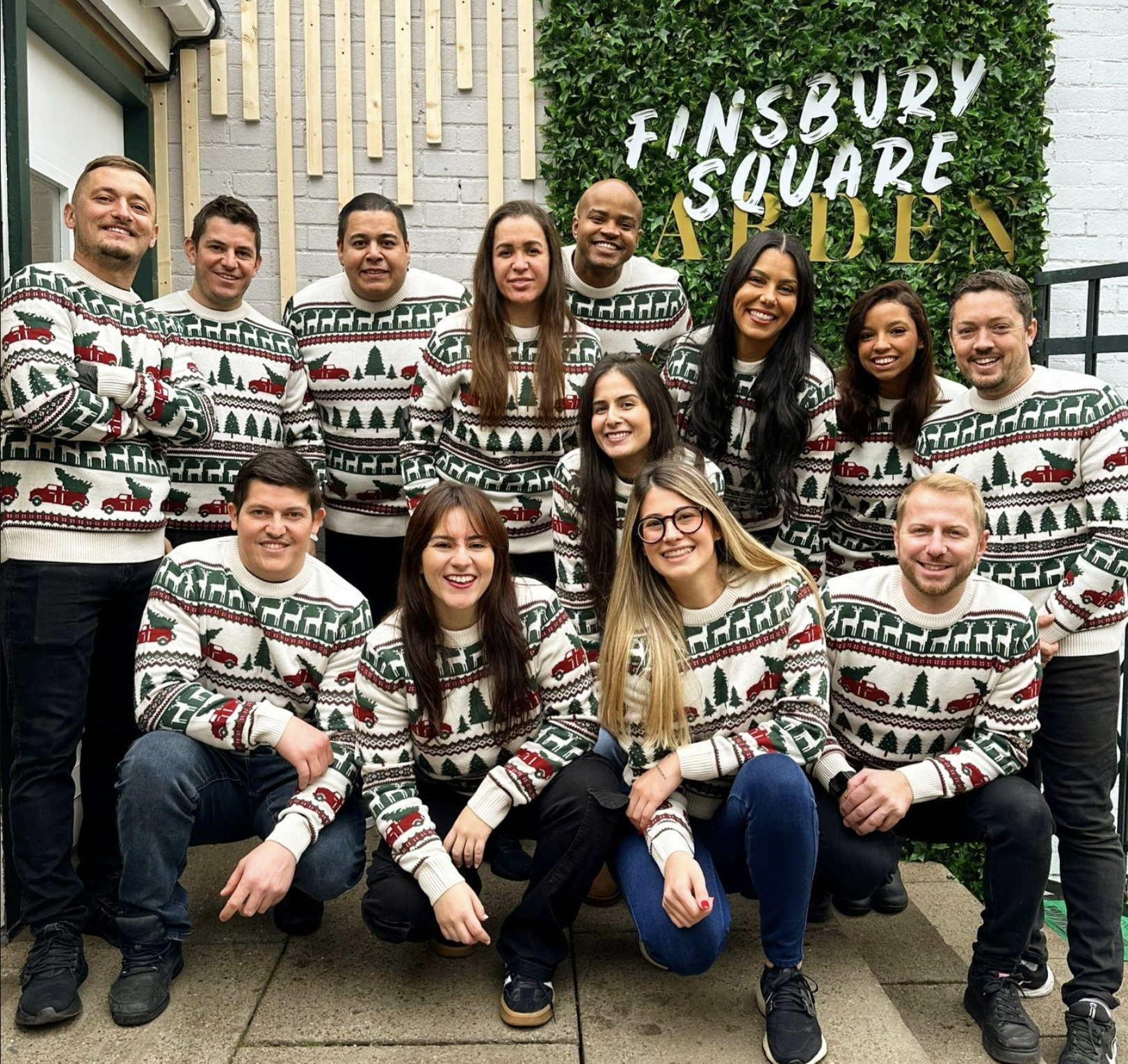 Group of people wearing Christmas sweaters, posing outside in front of a green wall with the sign "Finsbury Square" during festive season.