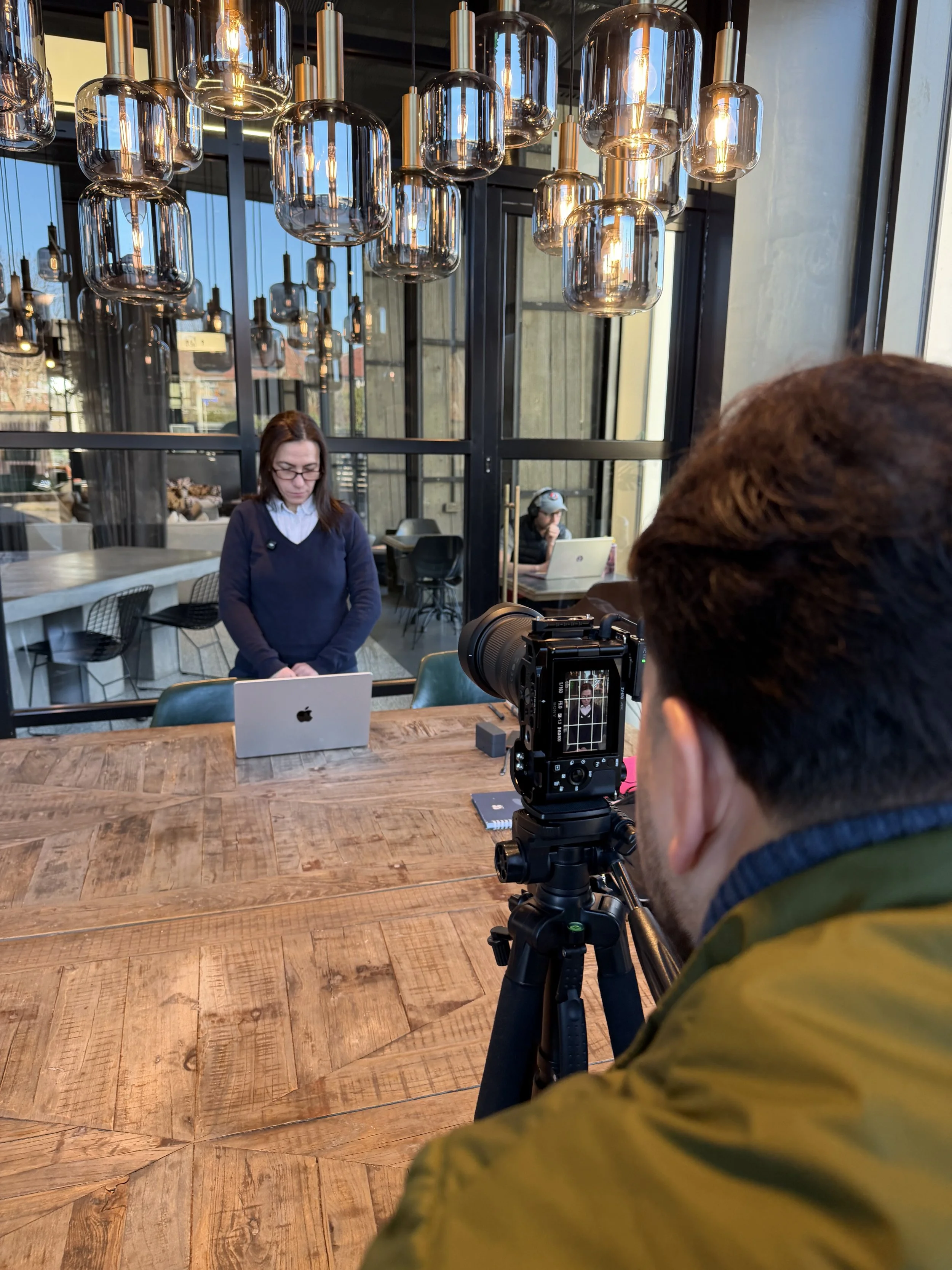 A person filming a woman in a navy sweater and glasses using a laptop in a modern cafe setting.
