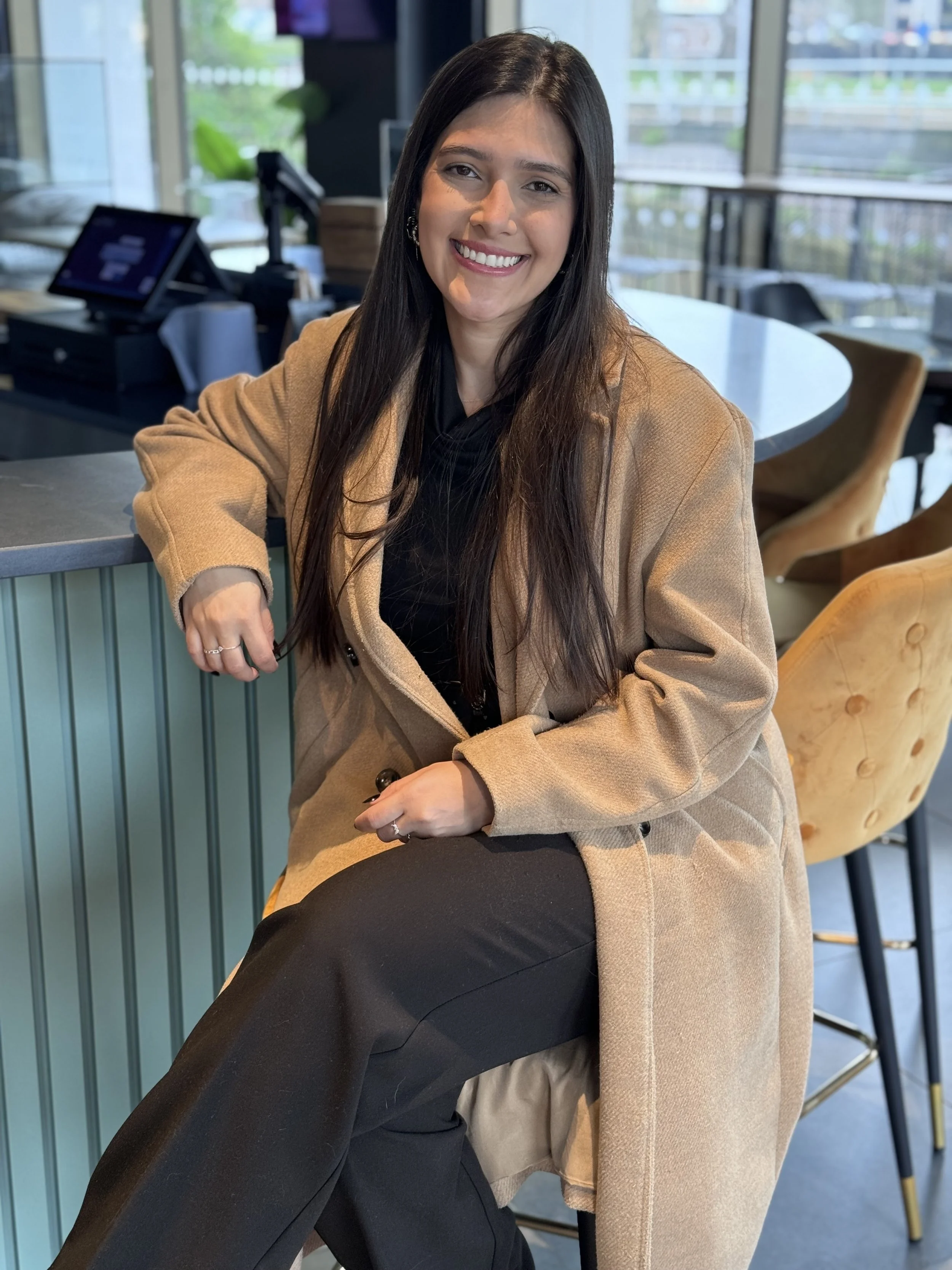 A woman with long dark hair, smiling, seated in a modern cafe with large windows, wearing a tan coat over black clothing.