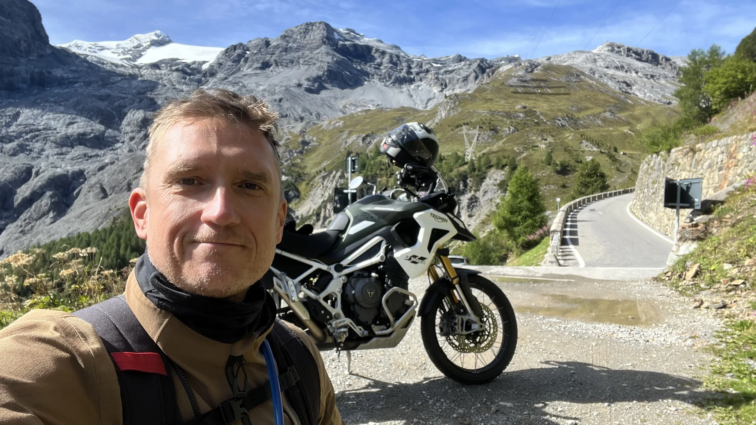 A man taking a selfie with a motorcycle parked on a mountain road, snowy peaks in the background, clear blue sky, and lush green trees.
