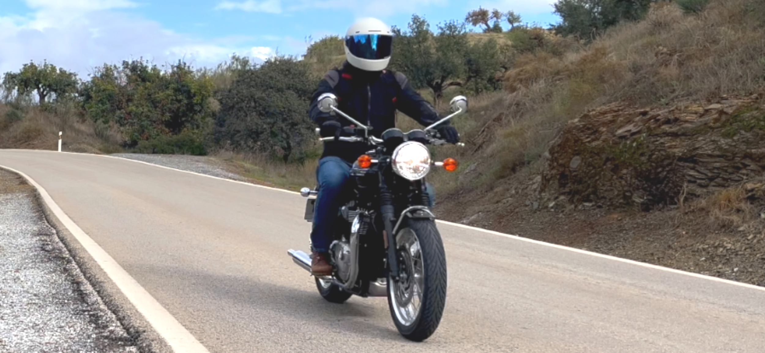 Motorcycle rider wearing a helmet and jacket riding on a winding road surrounded by rocky hills and trees.