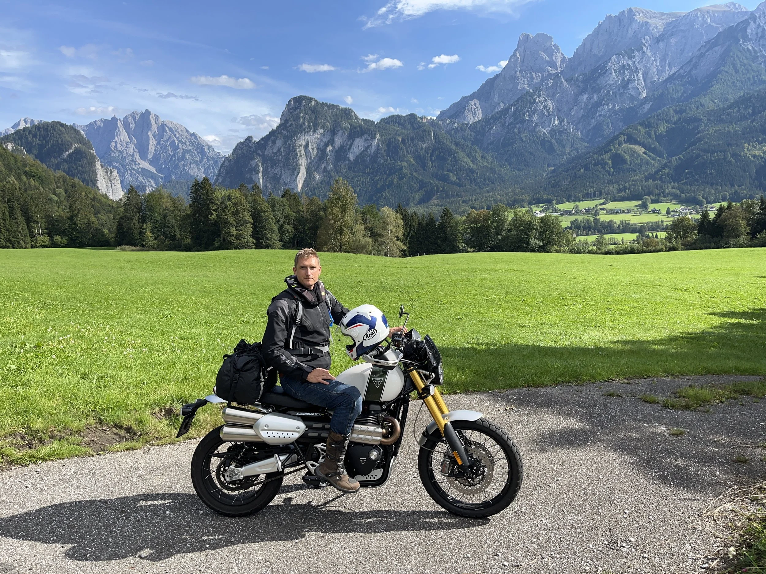 A man sitting on a white motorcycle with a mountain range and green fields in the background under a mostly cloudy sky.
