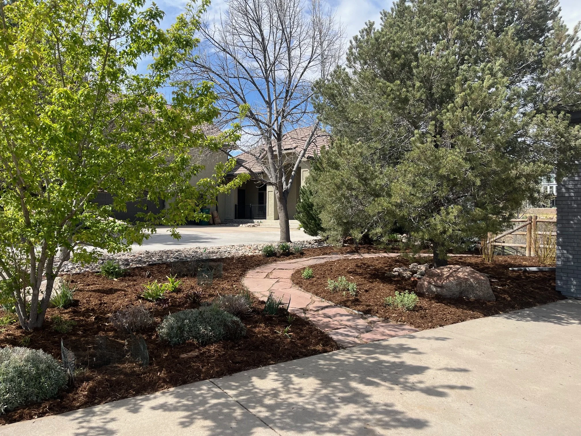 A residential front yard with a sidewalk, a stone pathway, trees, and shrubs. There is a house in the background with a tiled roof and a porch. The sky is partly cloudy.