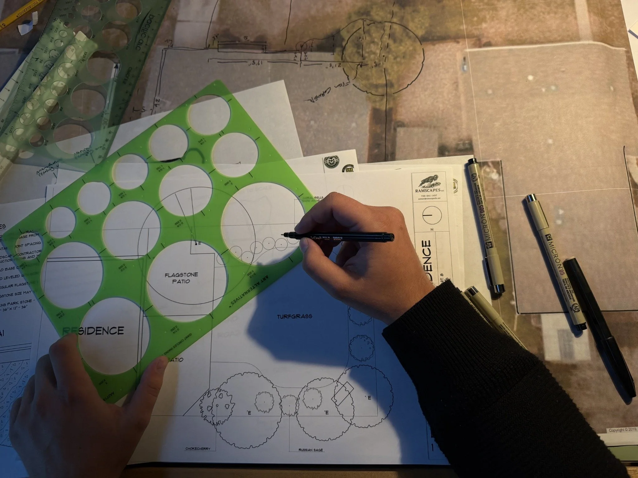 Architectural plans, layout diagrams, and drawing tools on a workspace, with a person's hand holding a pen.