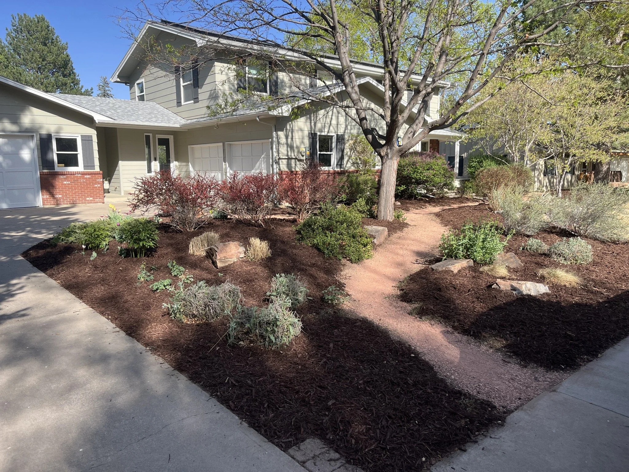 A house with a landscaped yard featuring a large tree, shrubs, and flowering plants, with partly cloudy skies in the background.