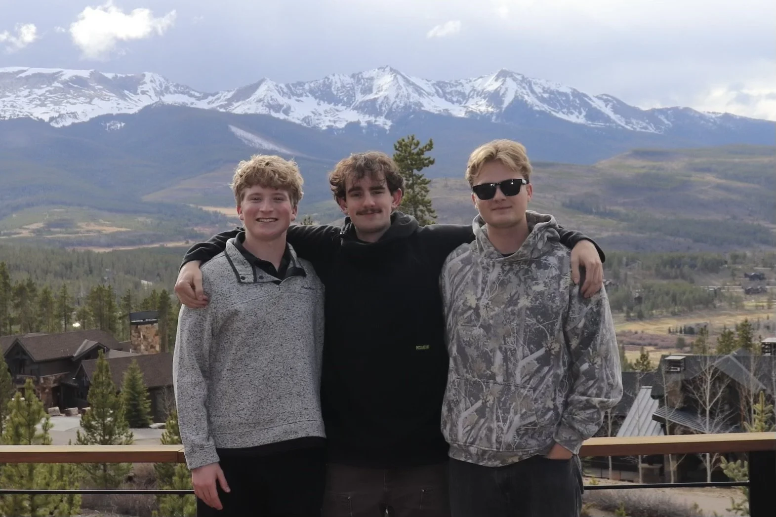 Three young men standing together outdoors with mountains, partially snow-capped, and a small town in the background. They have their arms around each other, smiling at the camera.
