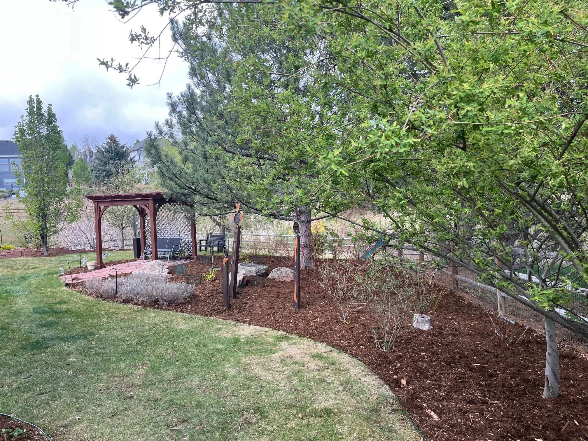 A backyard garden scene with a lush green lawn, a small raised garden bed surrounded by bricks, a wooden arbor, and various trees and plants. The background includes houses and a cloudy sky.