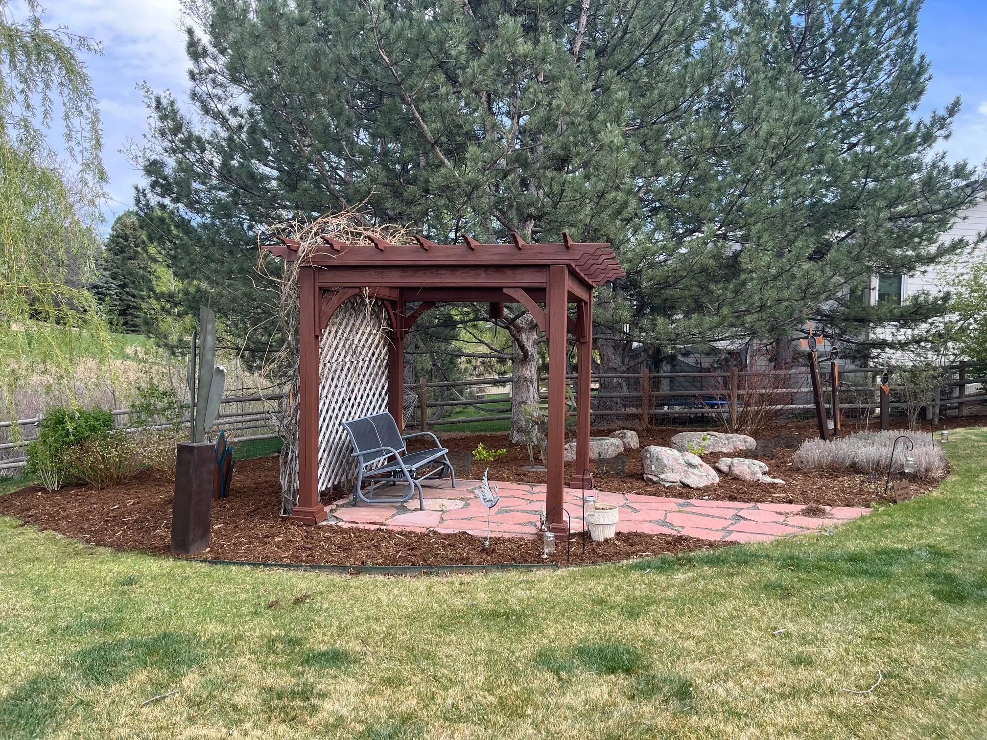 A backyard garden with a wooden arbor, two chairs, a stone pathway, and trees, including a large pine tree, under a partly cloudy sky.