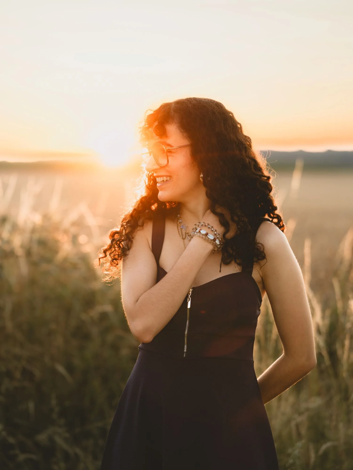 Jeune femme aux cheveux bouclés, portant une robe noire avec une fermeture éclair, souriant, en pleine nature au coucher du soleil.