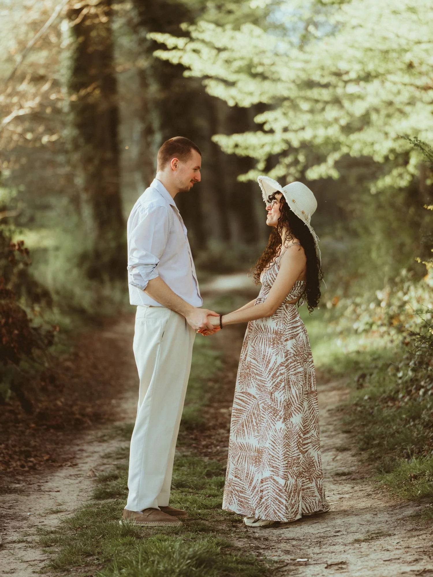Un couple se tient la main sur un chemin forestier, avec des arbres verdoyants en arrière-plan. L'homme porte une chemise blanche et un pantalon beige, tandis que la femme porte une robe longues imprimée de feuilles et un chapeau blanc. Ils se regardent en souriant.