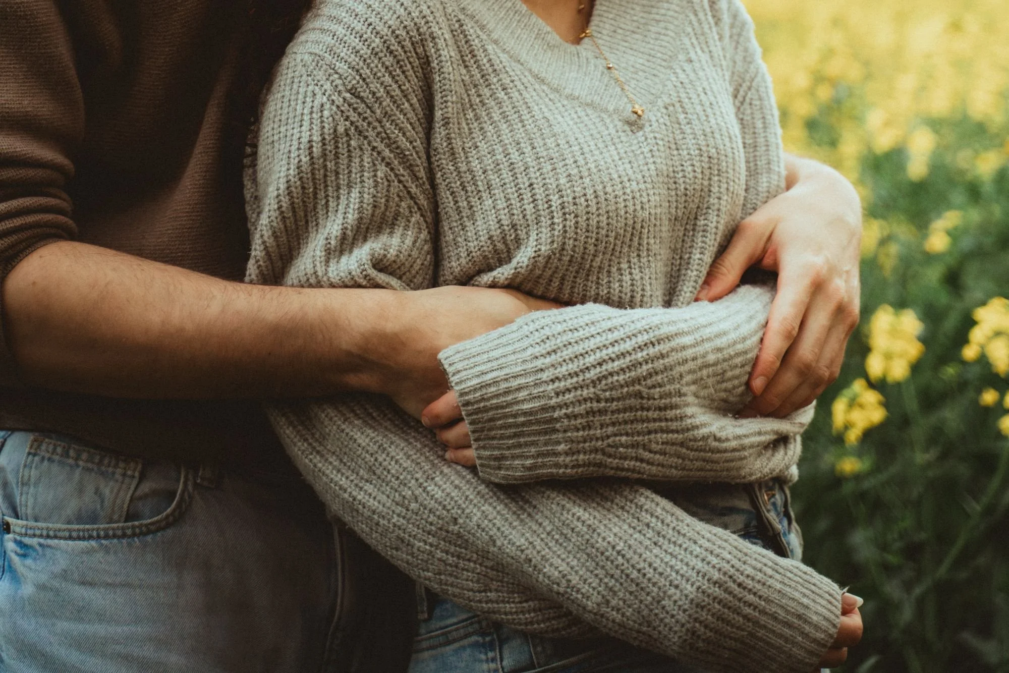 Deux personnes, dont une femme portant un pull en tricot beige, sont enlaçant dans une nature avec des fleurs jaunes.