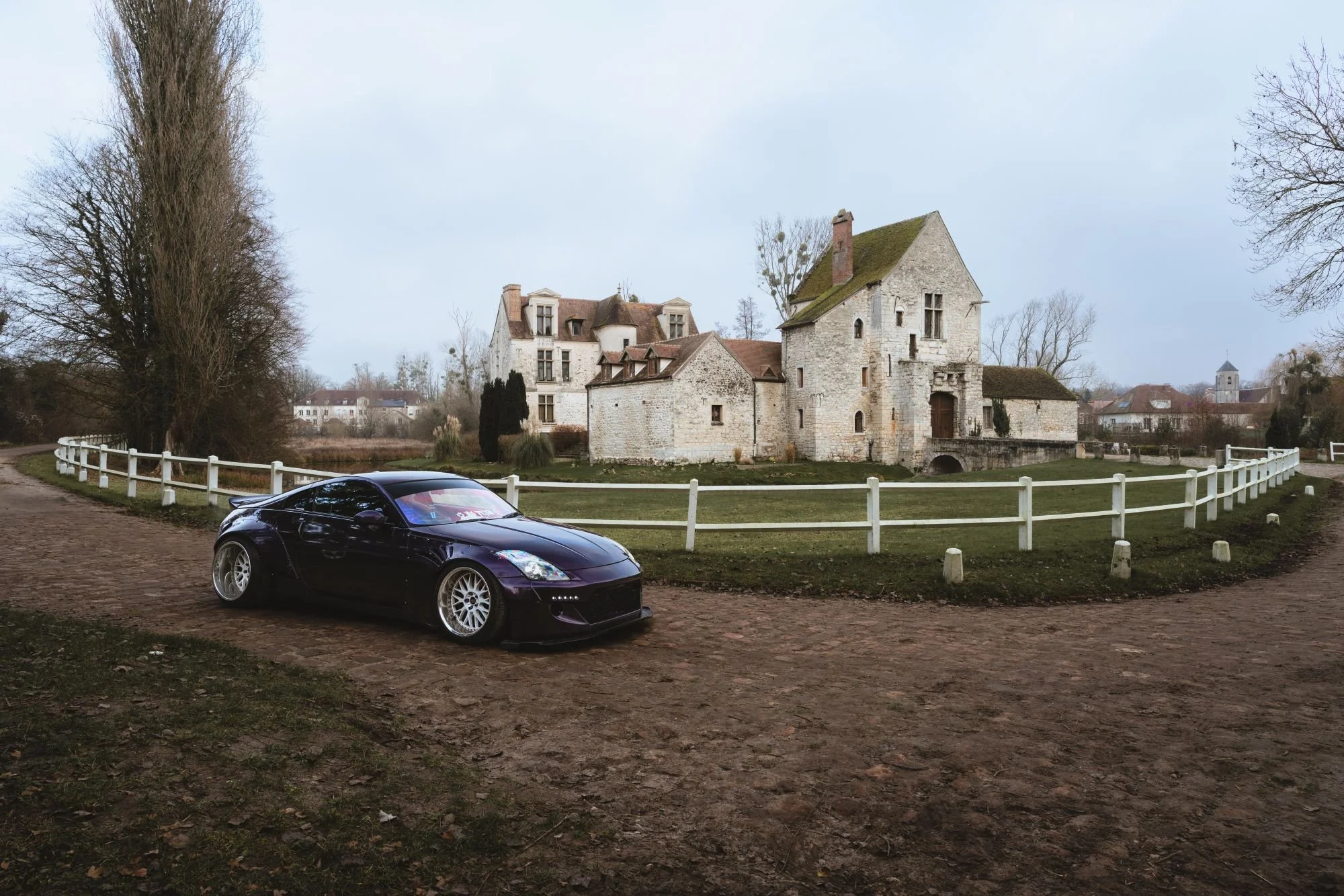 Une voiture noire moderne garée sur un chemin en terre devant un vieux château en pierre entouré d'une clôture blanche, avec des arbres dénudés et un ciel gris en arrière-plan.