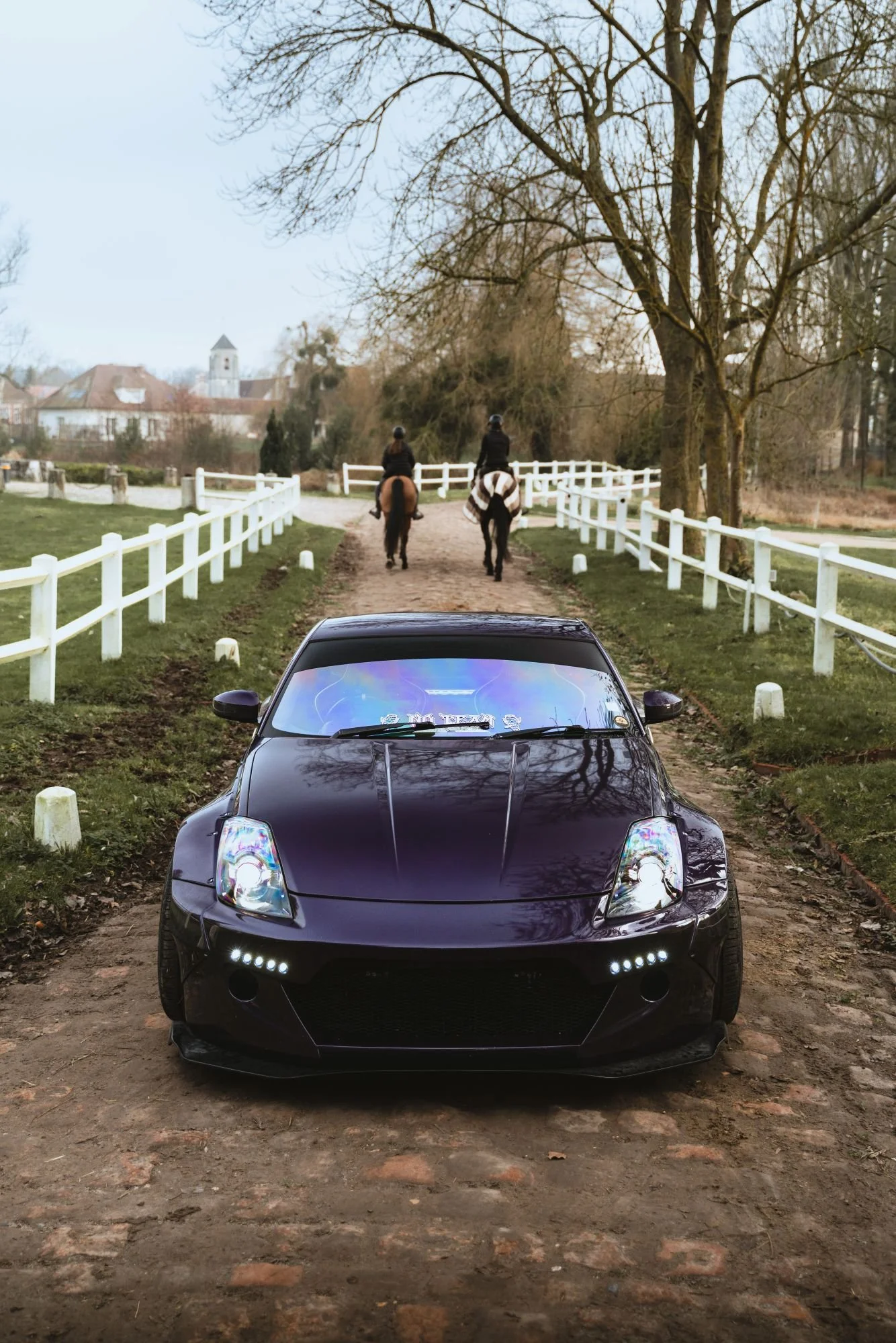 Voiture noire sur un chemin de terre, deux cavaliers à cheval à l'arrière, en milieu rural avec arbres dénudés et des maisons au fond.