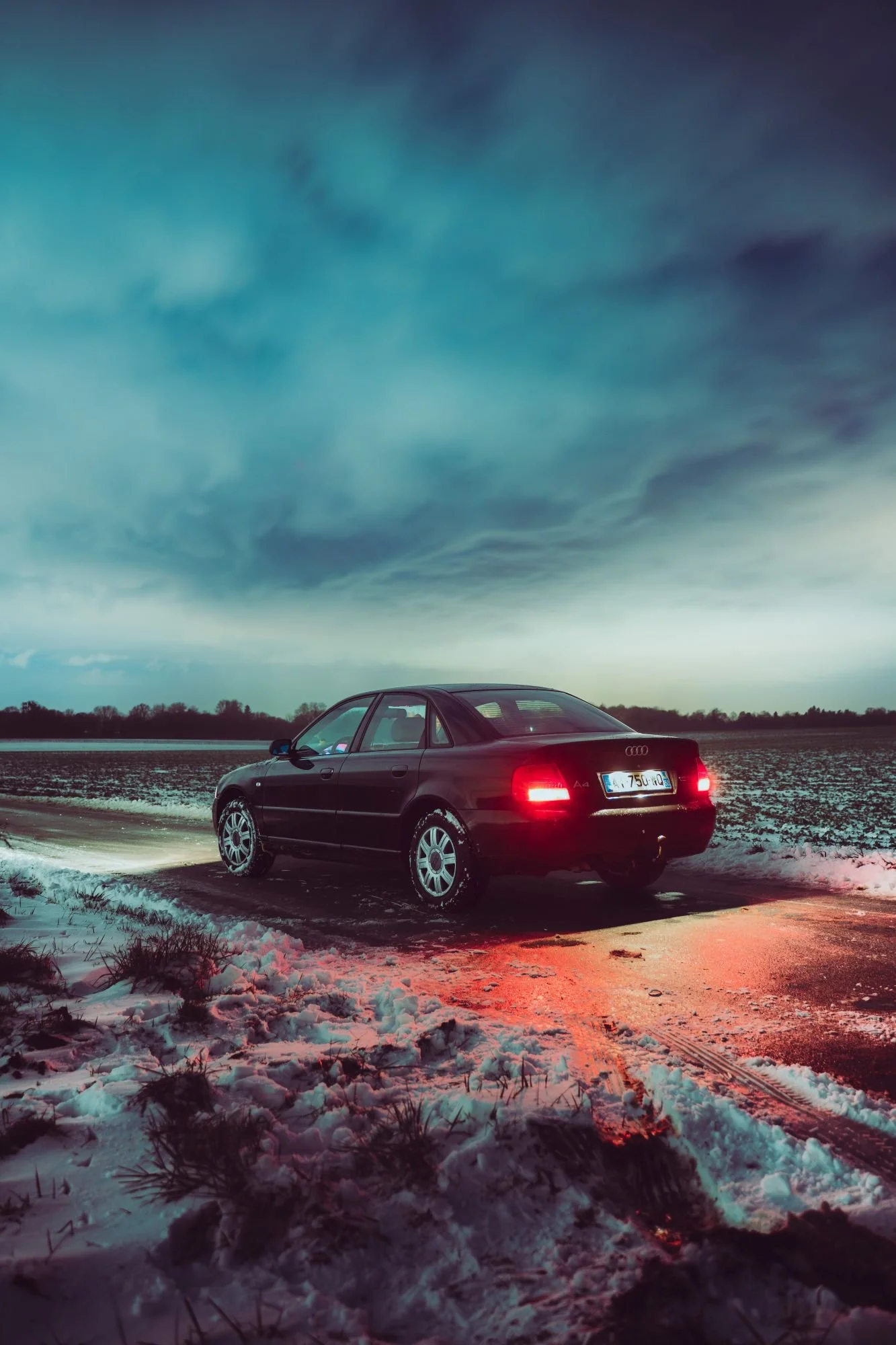 Une voiture noire Audi A4 stationnée sur une route enneigée lors d'une soirée nuageuse, avec des traces de pneus dans la neige et un ciel orageux en arrière-plan.