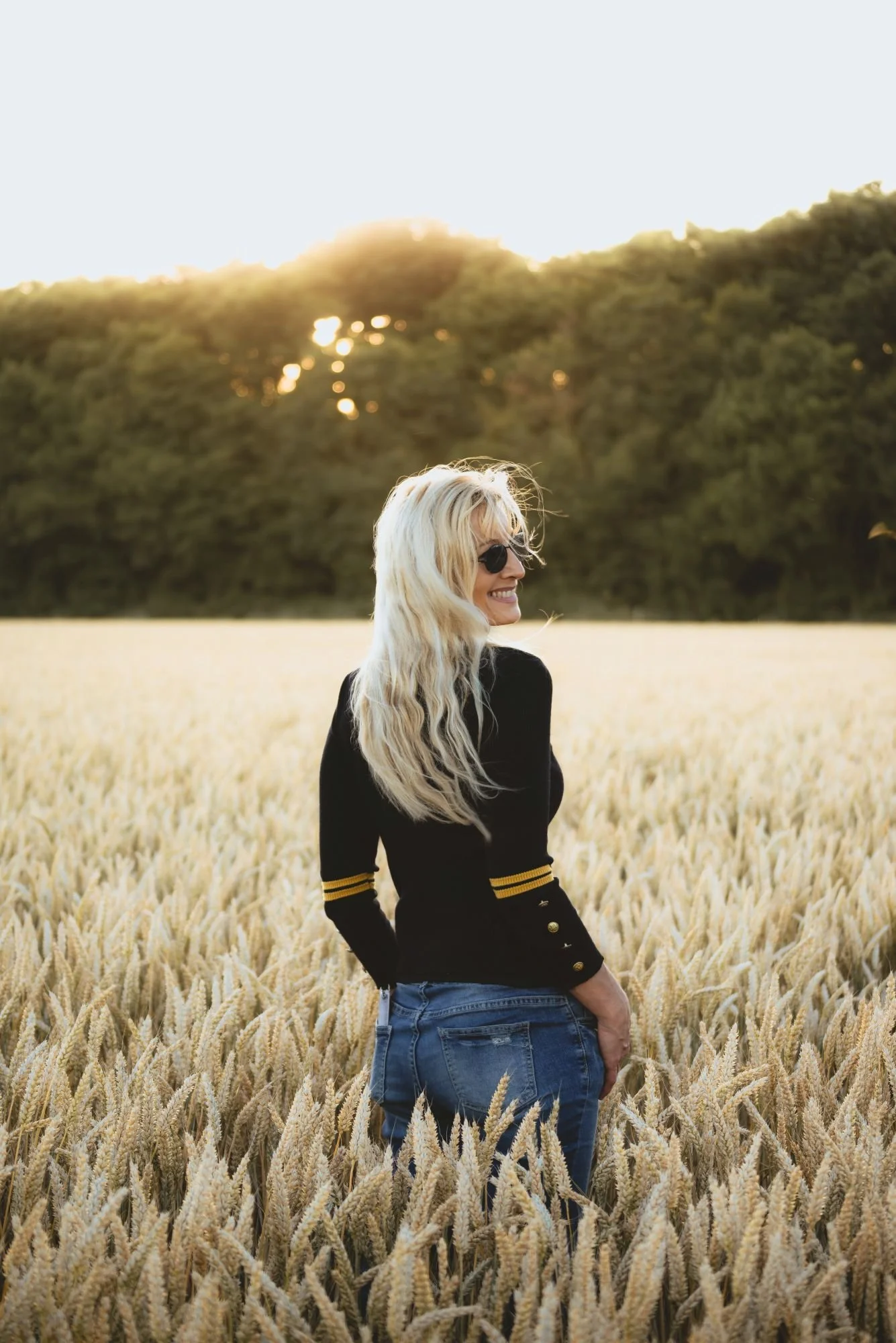 Femme souriante portant des lunettes de soleil, vêtue d'un pull noir avec des bandes jaunes sur les manches, dans un champ de blé au coucher du soleil.