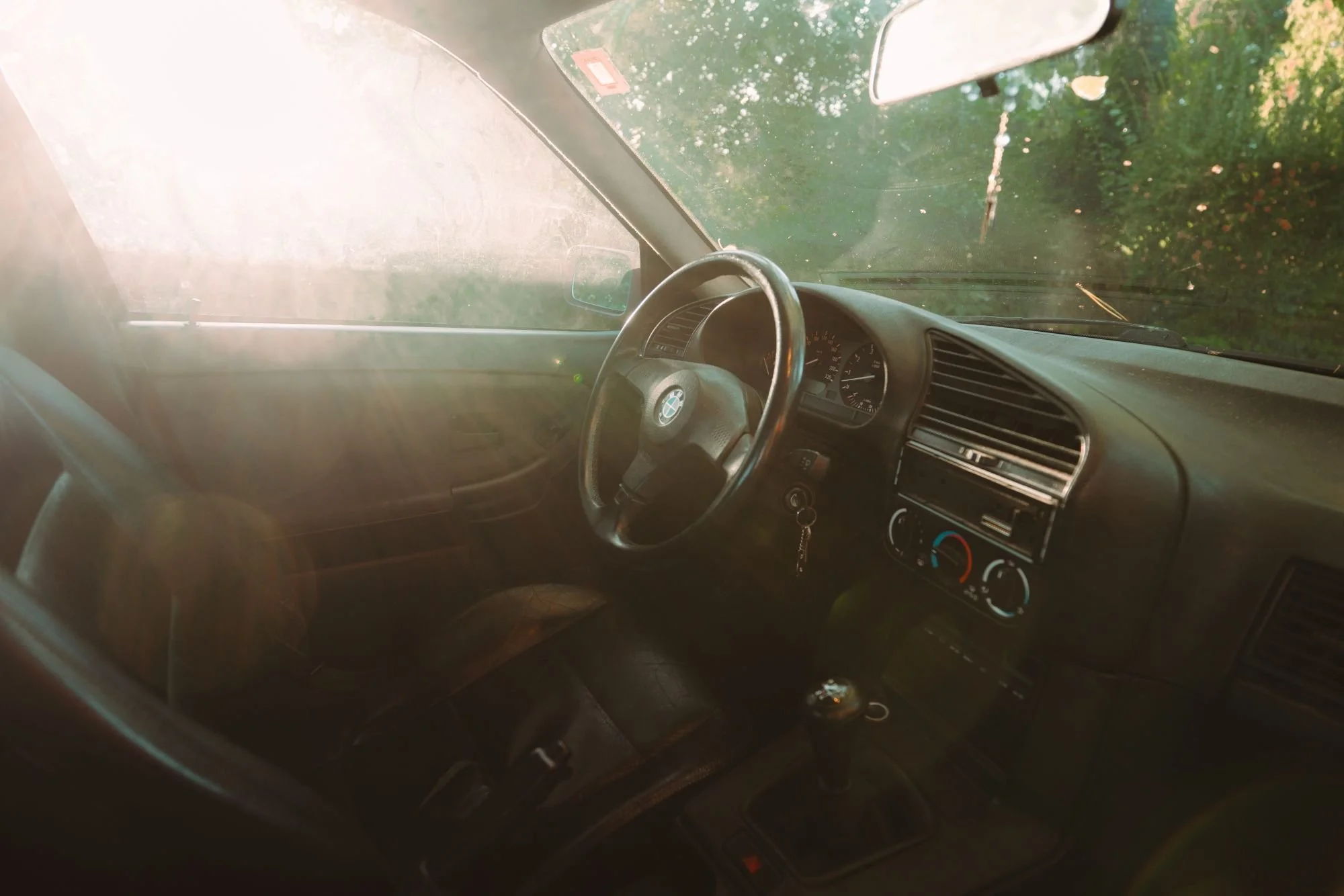 Intérieur d'une voiture vue de l'avant, avec le volant, le tableau de bord, les commandes de climatisation, et une vue vers la fenêtre montrant la végétation extérieure