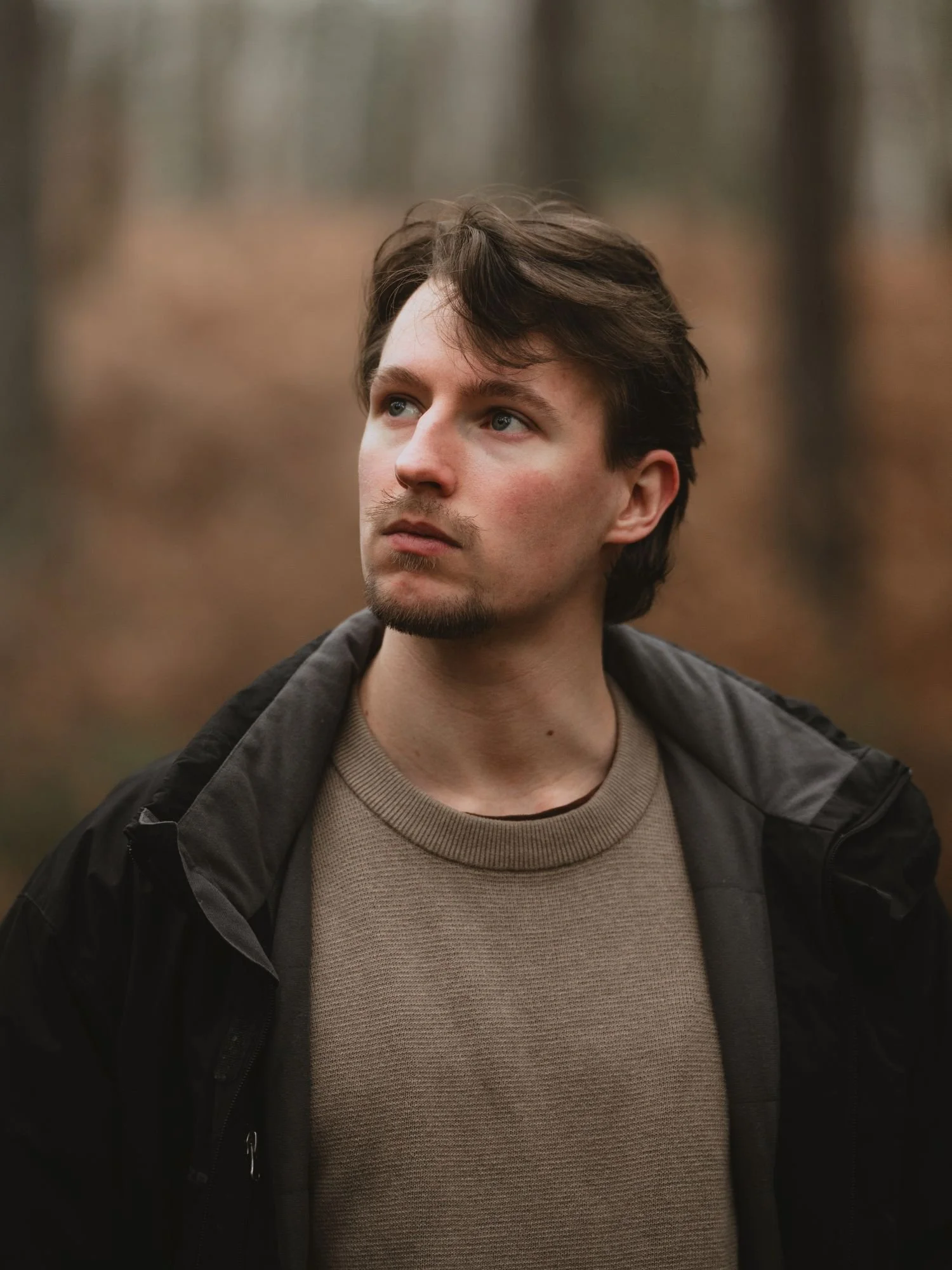 Un jeune homme avec des cheveux bruns et des yeux bleus, portant un manteau noir et un pull marron, se tient dans une forêt aux couleurs automnales.